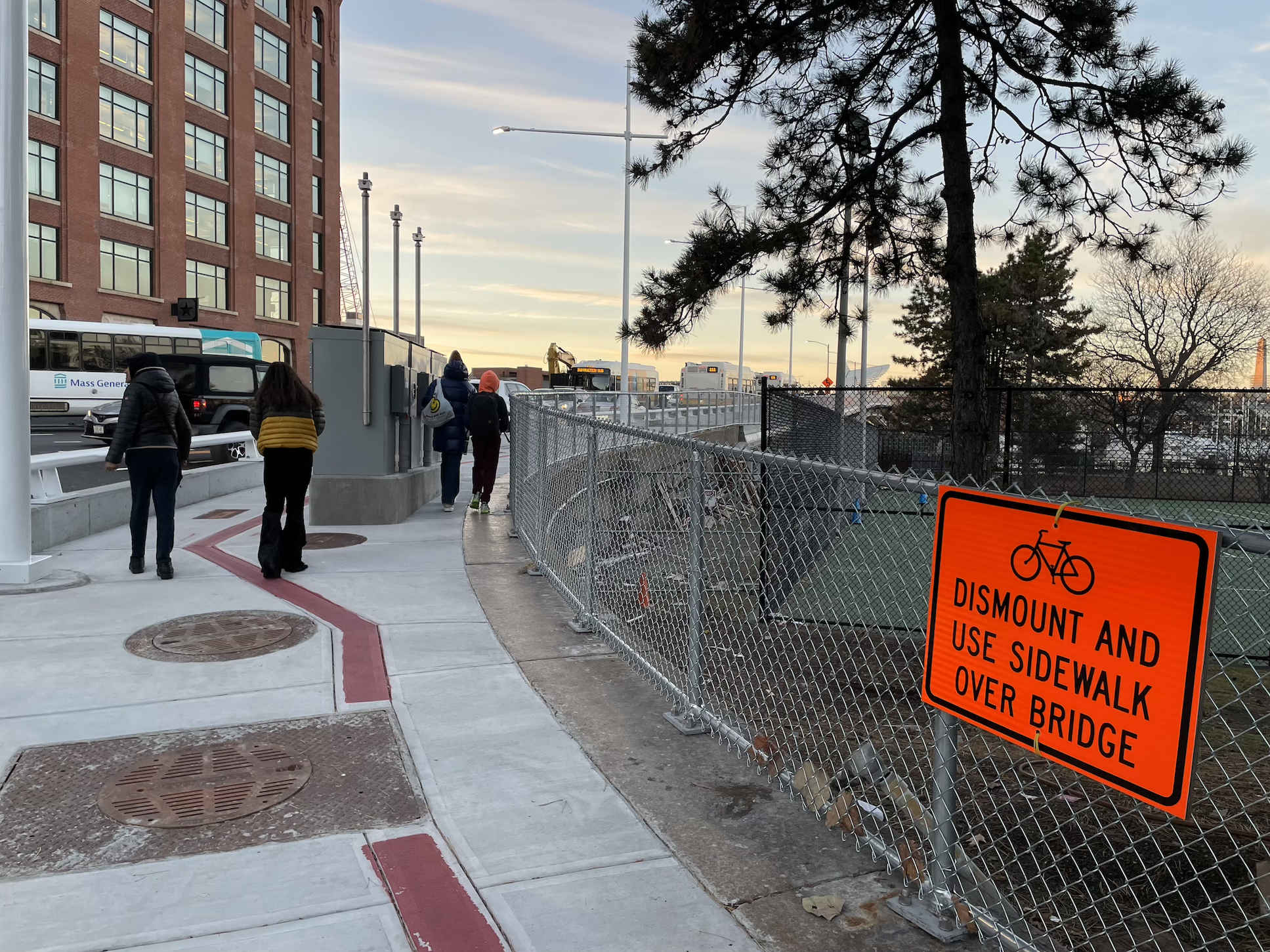 An orange sign attached to a chain-link fence reads "DISMOUNT AND USE SIDEWALK OVER BRIDGE" under a bike icon. To its left, several pedestrians walk on a sidewalk towards a bridge. In the middle of the sidewalk is a red stripe that denotes the Freedom Trail. Several pine trees are on the other side of the chain-link fence.