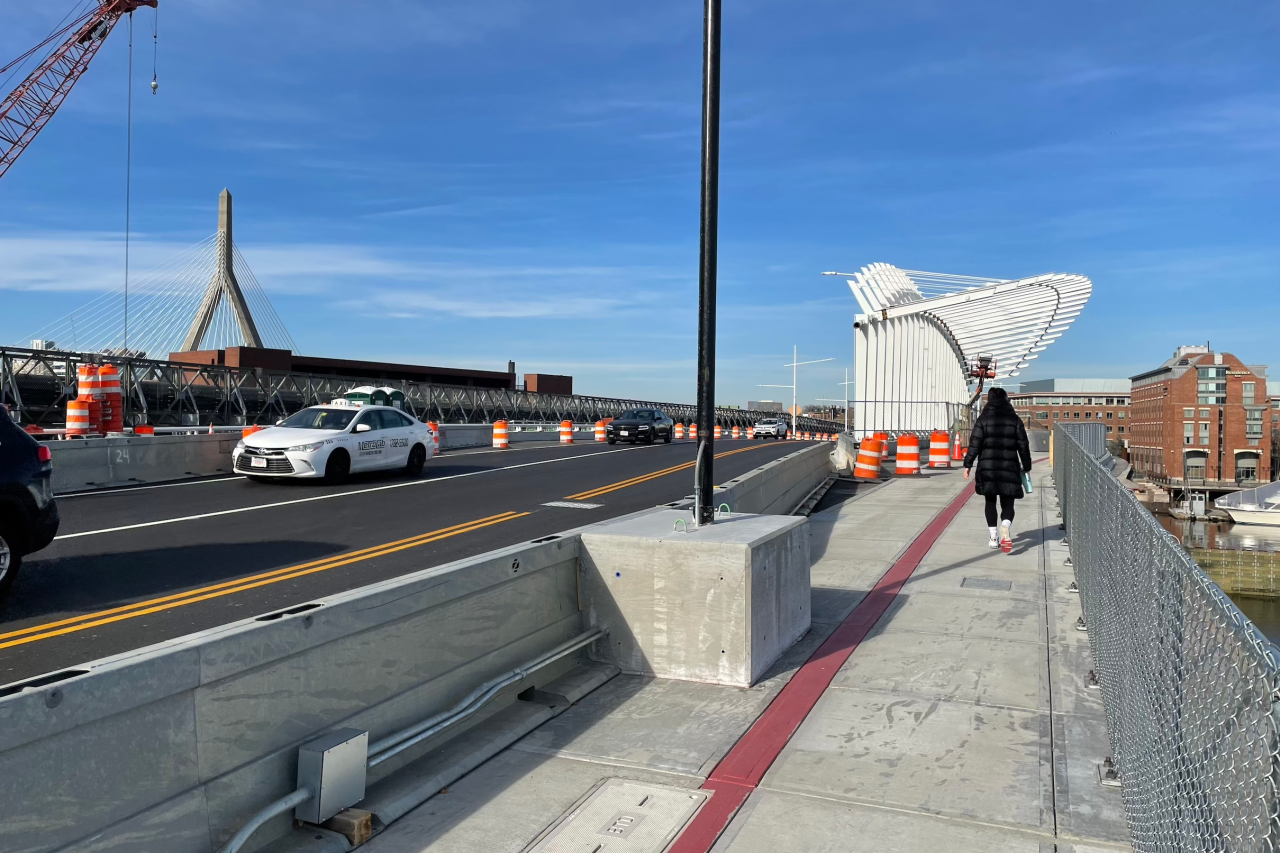 A pedestrian walks on a sidewalk next to some temporary crash barriers on a new bridge. To the left several cars drive on fresh asphalt, and on the left edge a crane is visible among some orange construction barrels.