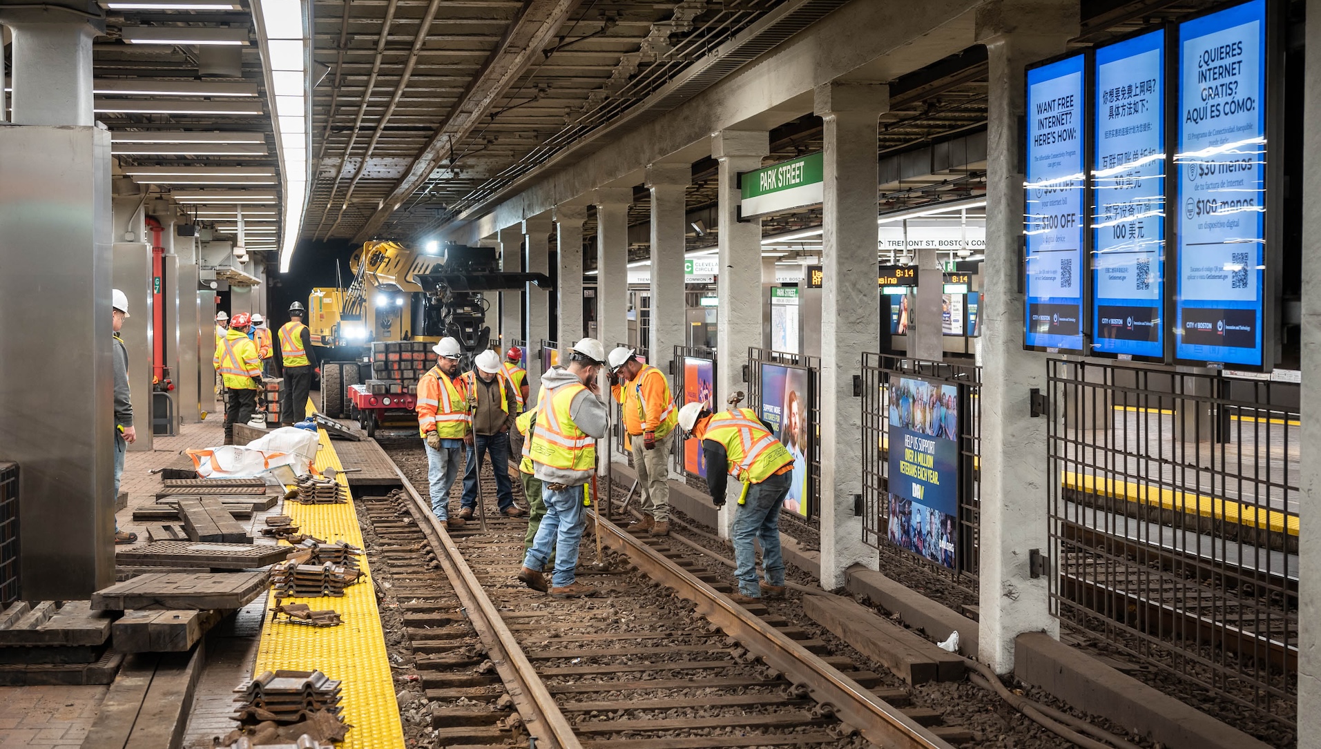 Workers in fluorescent yellow vests and helmets place tracks inside a subway station below a green "Park Street" sign. The platform of the station is covered in railroad ties and plates and other construction materials. In the distance a yellow lull forklift is on the tracks at the edge of the station.