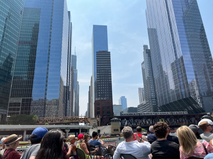 A Chicago city skyline full of skyscrapers with glass windows with a subway passing by down below and boat passengers looking on.
