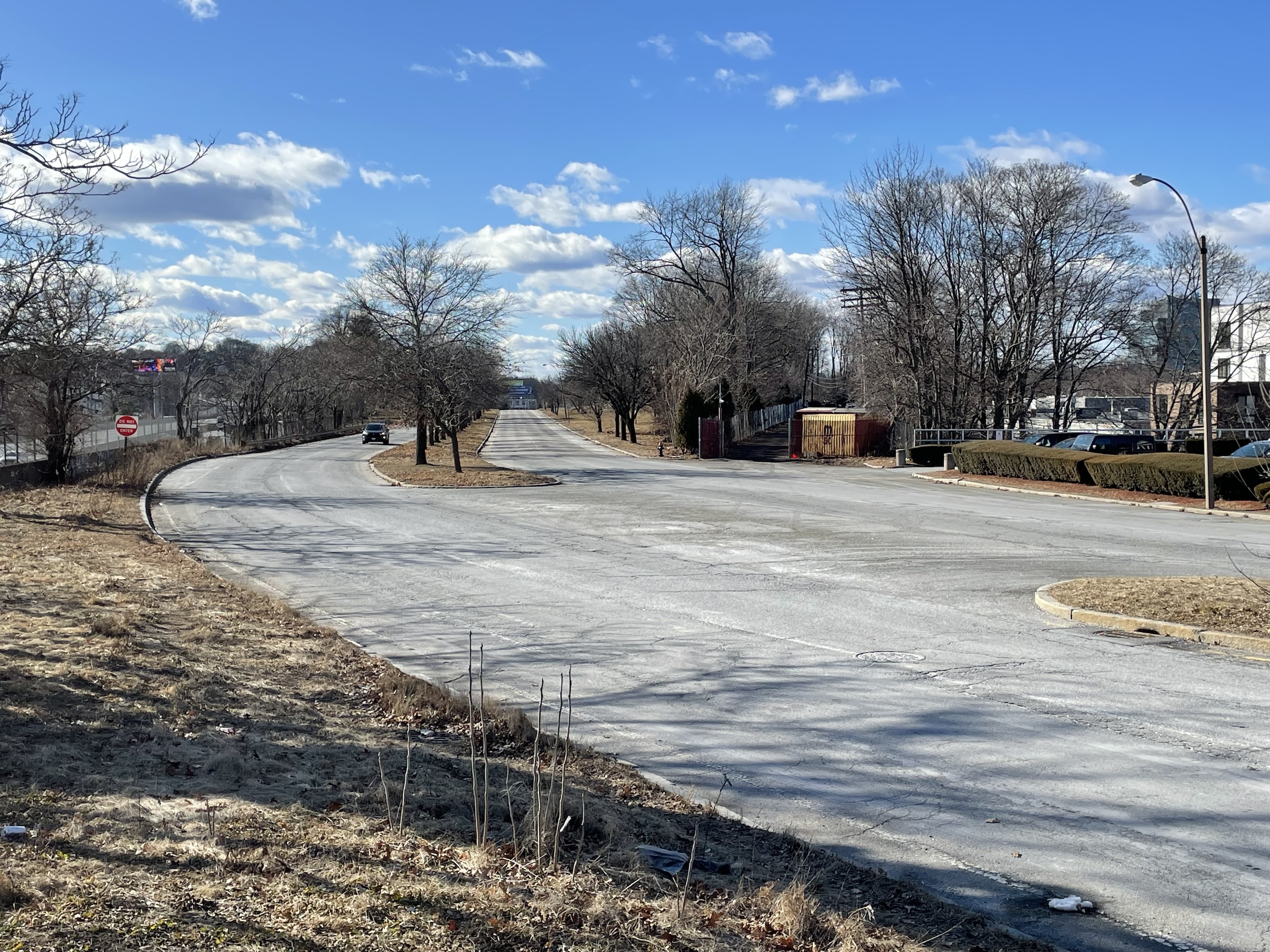 A long, empty four-lane highway bisected by.a median with trees stretches toward the horizon. A single car approaches in the distance.