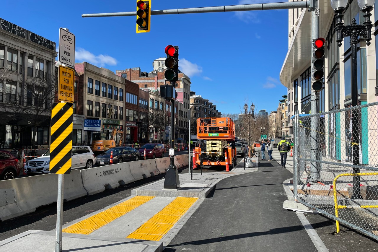 A new sidewalk, new traffic lights, and a protected bike lane run along a construction site. On the opposite side of the street are several older multi-story brick buildings.