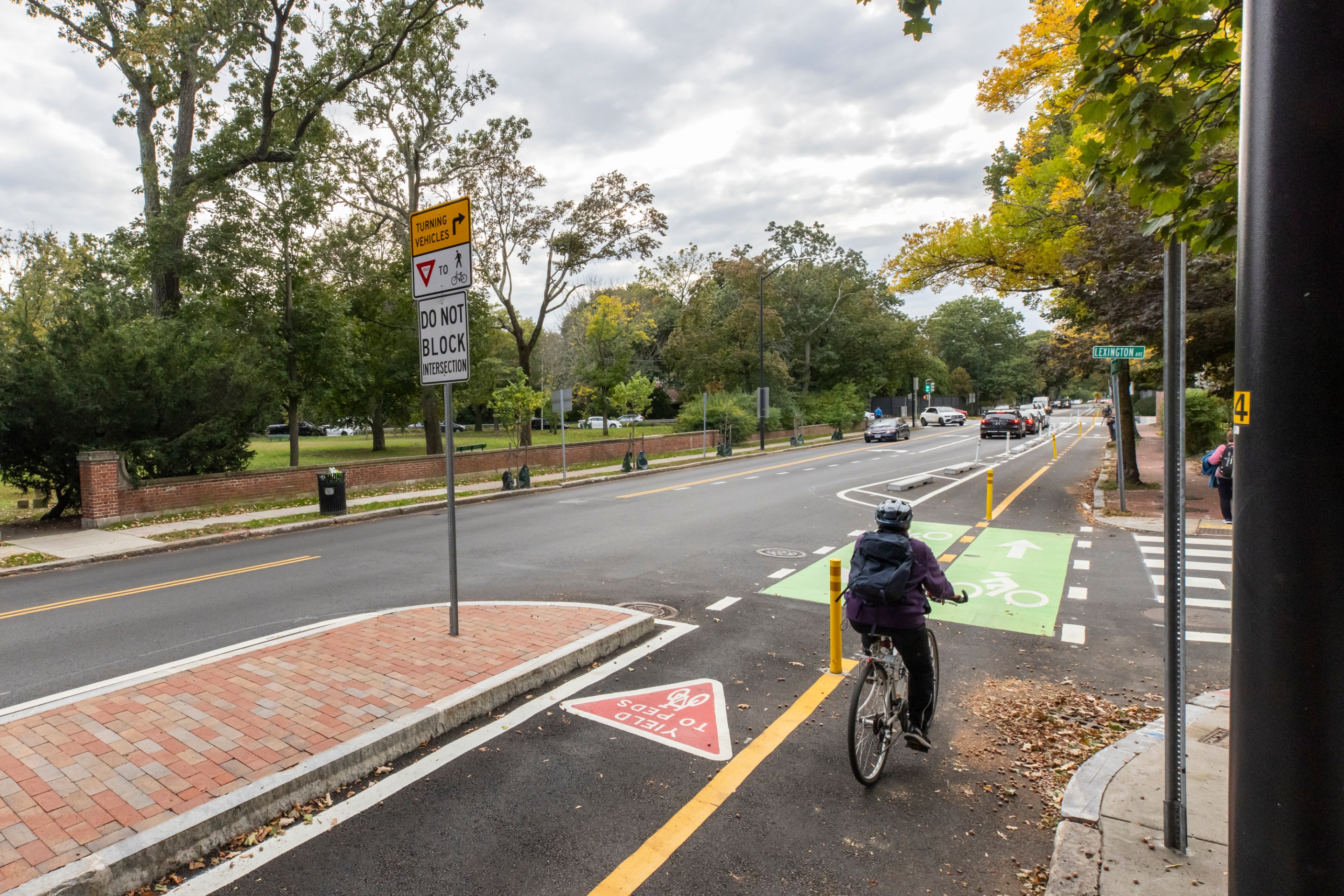 A person wearing black pedals a bike away from the camera on a wide street lined with trees beginning to turn yellow in the autumn. The rider is using a protected bike path that's separated from the adjacent car lanes by barriers including a brick-paved median island in the lower left foreground.