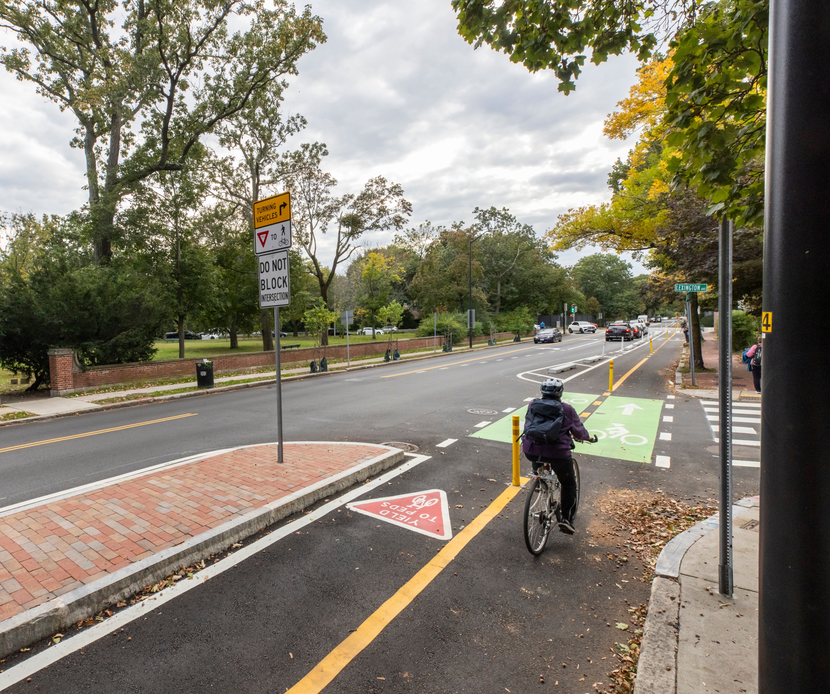 A person wearing black pedals a bike away from the camera on a wide street lined with trees beginning to turn yellow in the autumn. The rider is using a protected bike path that's separated from the adjacent car lanes by barriers including a brick-paved median island in the lower left foreground.