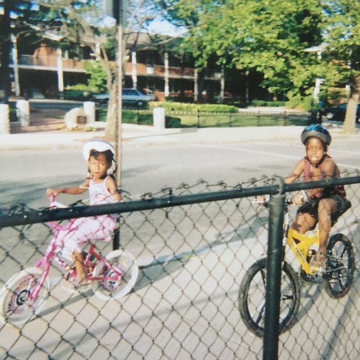 Two young Black children riding bikes behind a chain link fence.