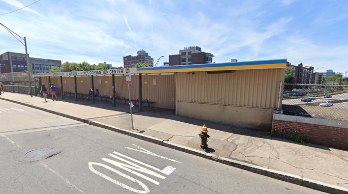 A beige metal bus shelter and broken sidewalk. Behind the bus shelter the Massachusetts Turnpike is visible. Courtesy of Google.