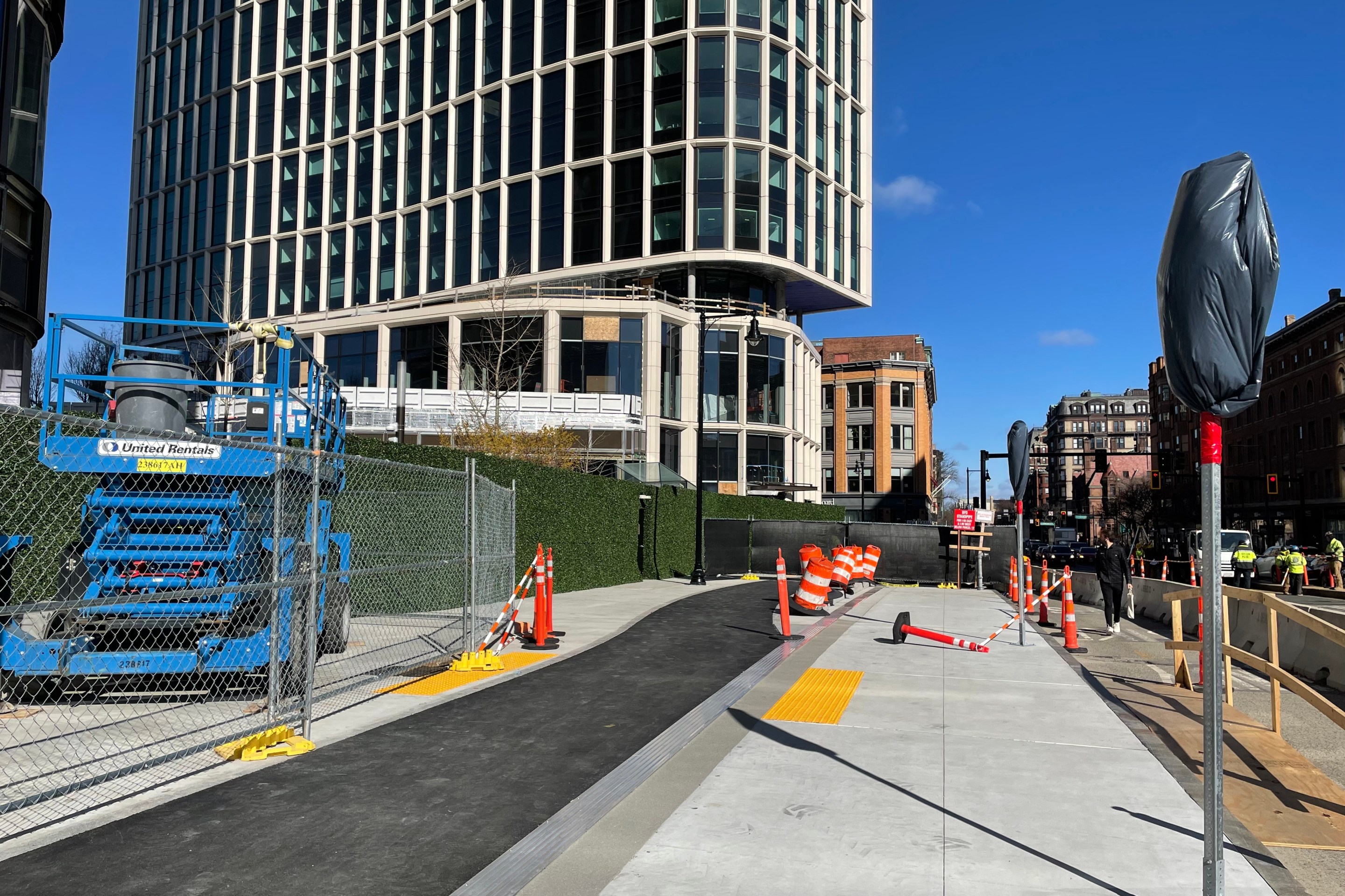 A sidewalk under construction with as asphalt bike path running down the middle. In the distance a new high-rise building rises behind a construction fence.