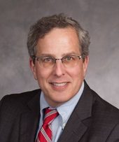 A white man with greying brown hair wearing a suit and red tie.