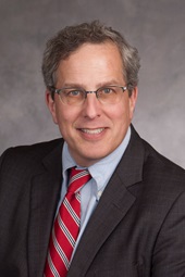 A white man with greying brown hair wearing a suit and red tie.