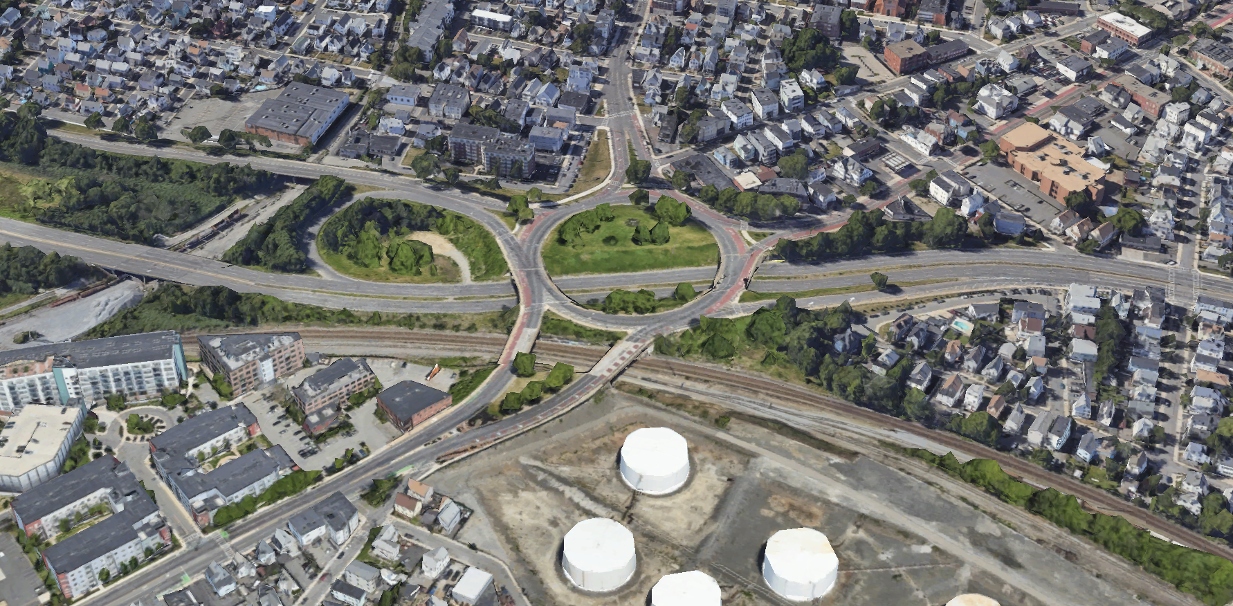 An aerial view of a large traffic rotary on a major highway that runs left-right through the center of the image. On the bottom edge are several large oil tanks and some railroad tracks. On the upper half of the image are densely-populated residential and commercial neighborhoods.