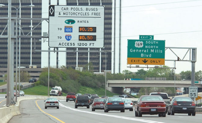 A picture of a wide interstate highway with two high-rises in the background. A white highway sign above the left lanes says "Car pools, buses & motorcycles free" and below that shows toll rates in an electronic sign. The toll is $0.25 for trips to Route 100 and $0.50 for trips to I-94.