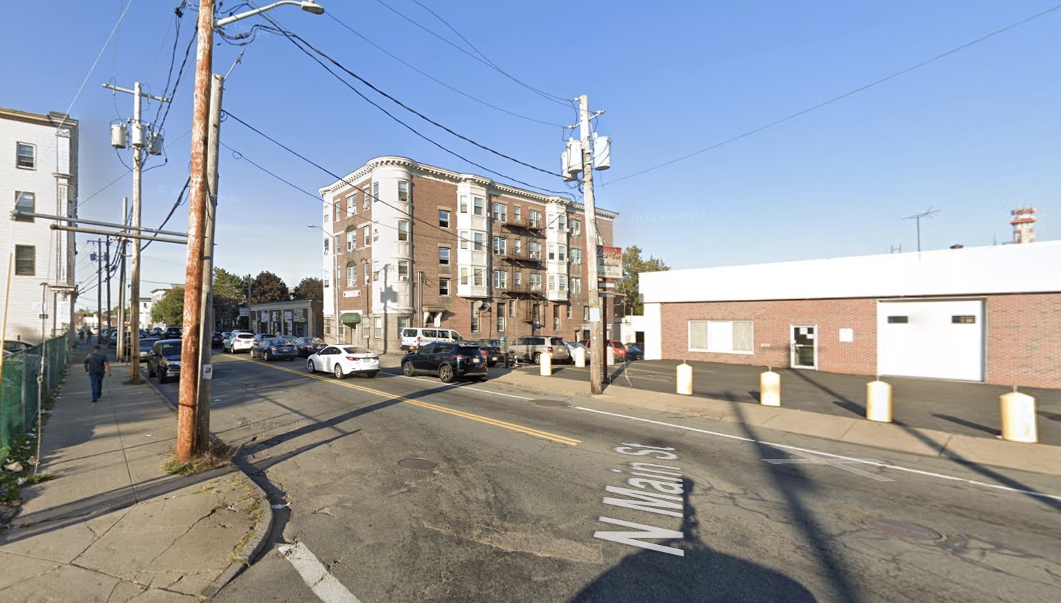 A Google Street View image of a street corner with several multi-story apartment buildings in the distance. In the foreground is a single-story commercial building set back from the street behind a parking lot.