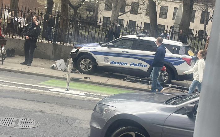 A cellphone snapshot of a crash scene where a Boston Police cruiser is on a sidewalk, with the wreckage of a traffic light underneath it. In front of the cruiser, near the left edge of the photo, a white man in a police uniform talks on a cellphone. Several pedestrians are walking around the crash site by walking in the street in the foreground. Behind the crashed car is a iron fence surrounding a graveyard where numerous people in coats are walking.