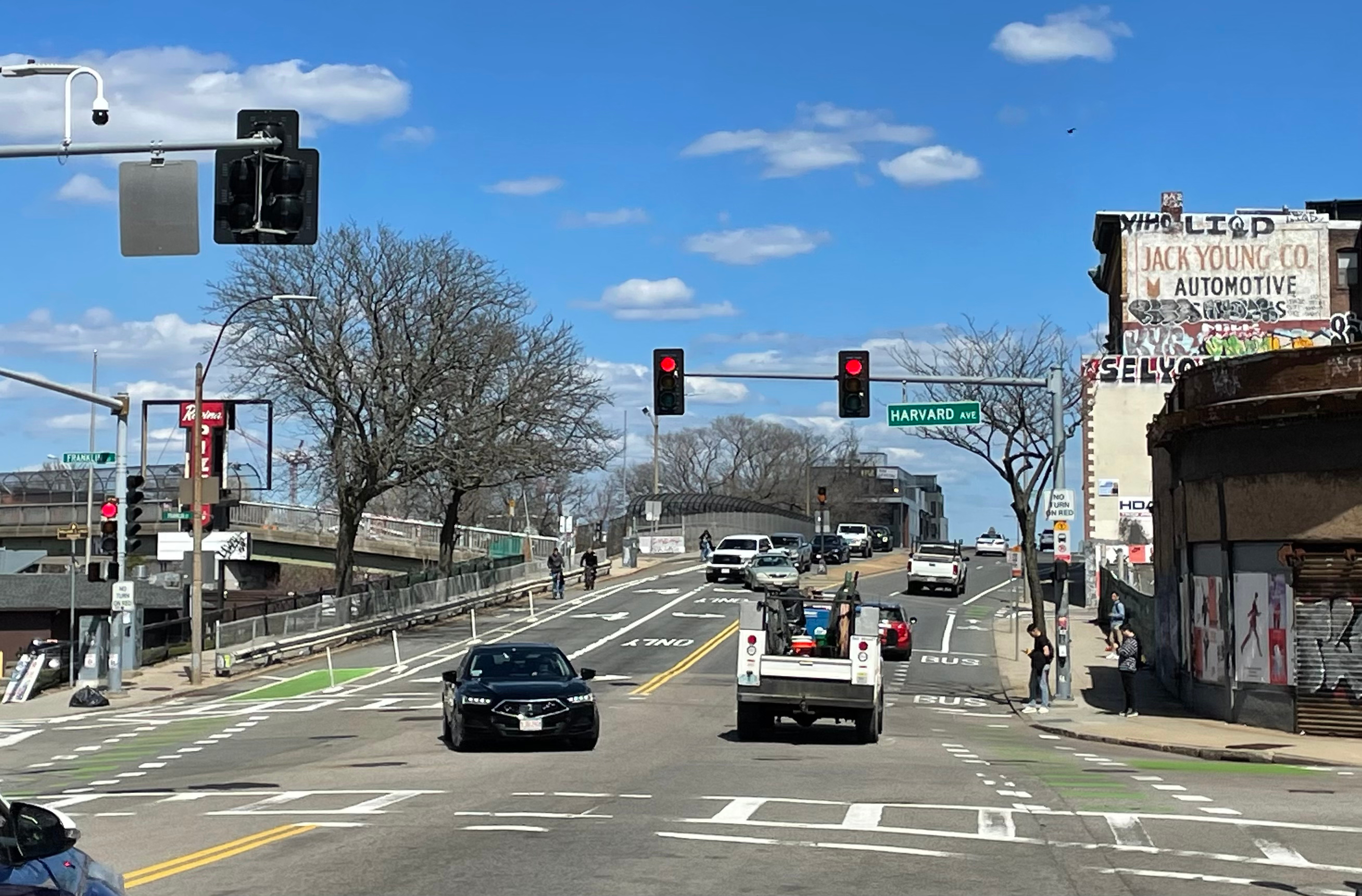 A view up a city street under blue skies. A street sign on a traffic light reads "Harvard Ave." Several people on bikes ride towards the camera in a bike lane near the left side of the photo.