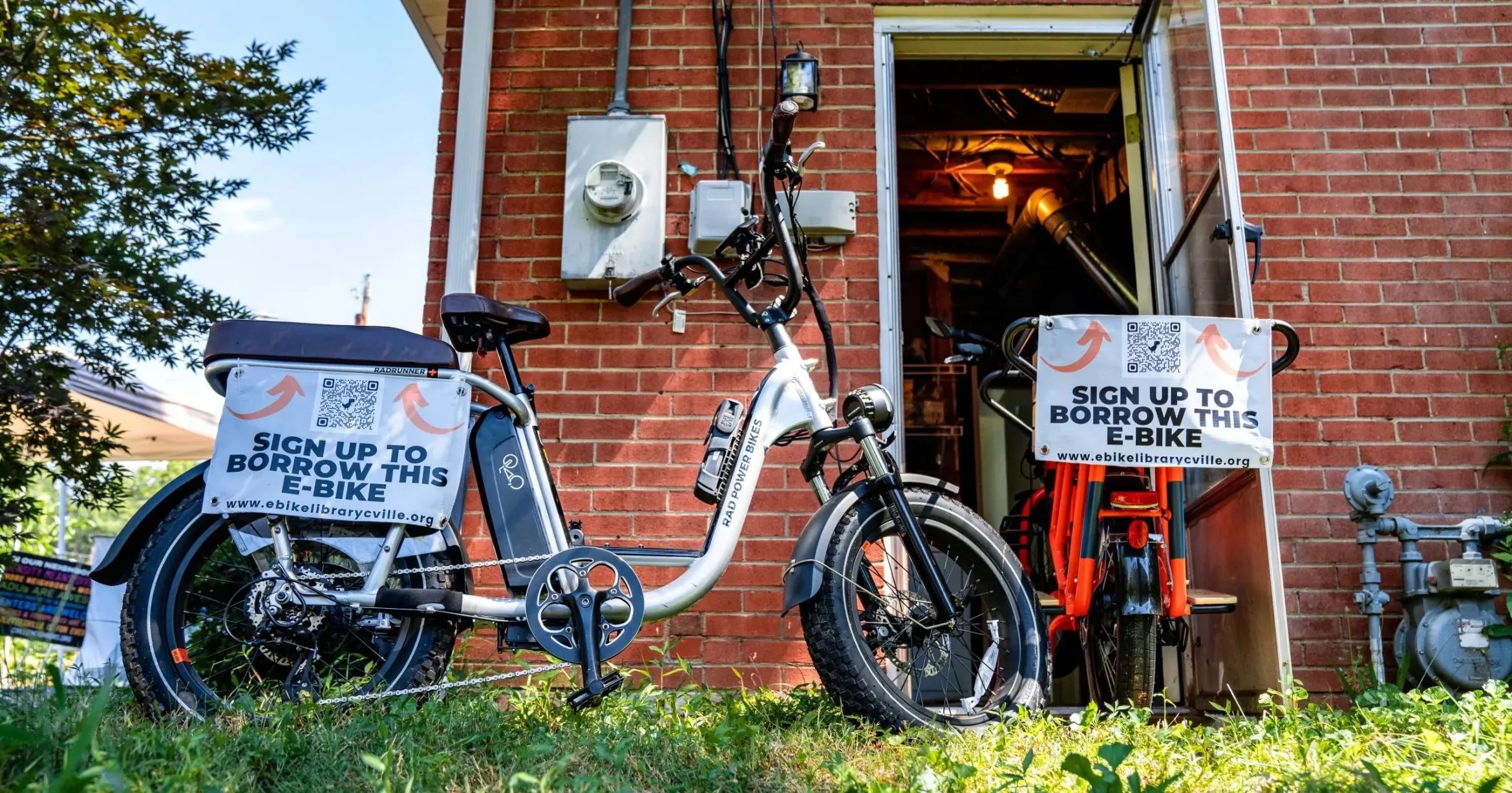 Two bikes parked by a shed with signs that say "sign up to borrow this e-bike" with a QR code.
