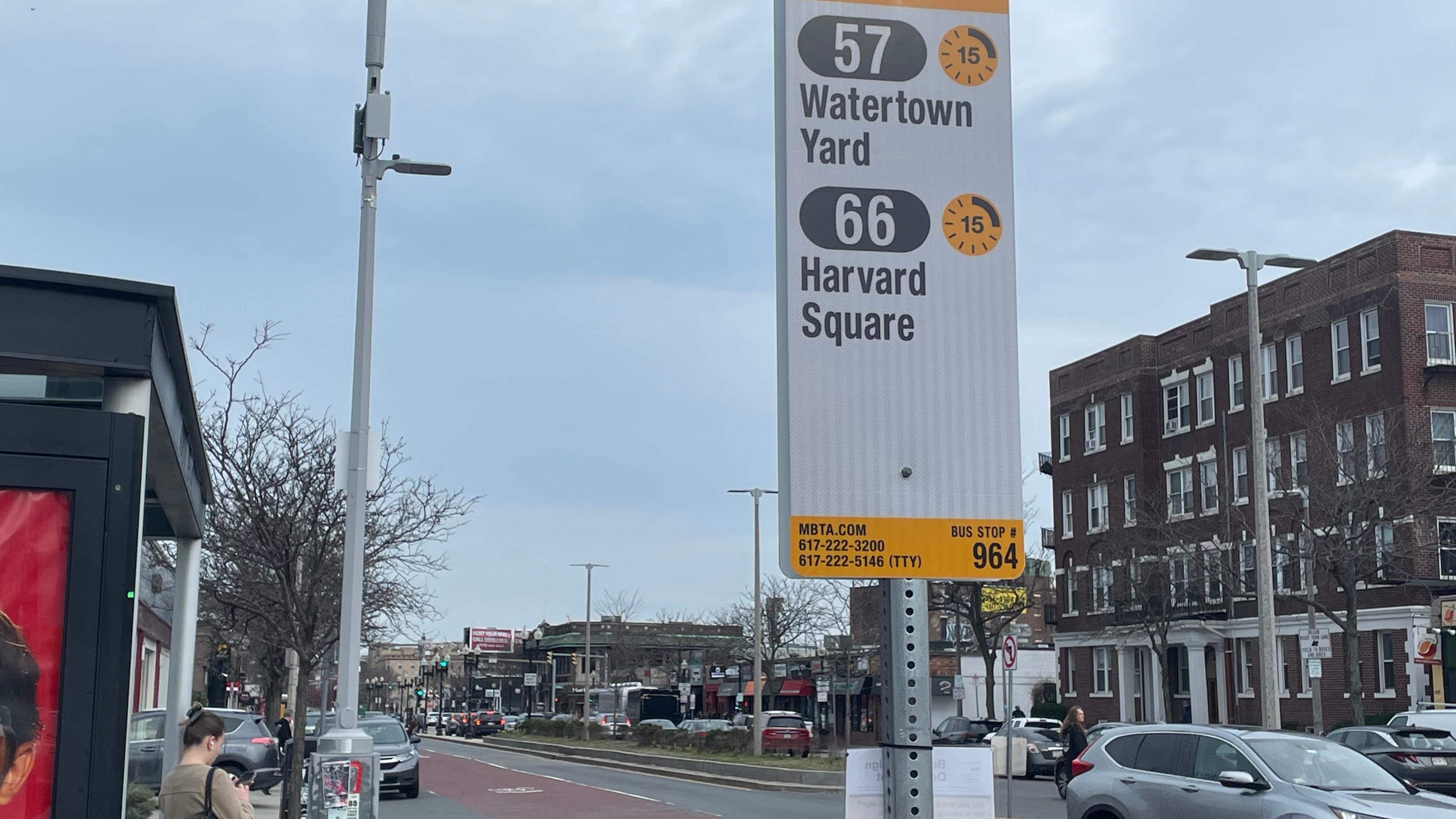 A bus stop sign next to a bus shelter where a passenger is waiting shows two bus routes - the 75 to Watertown Yard and the 66 to Harvard Square - next to a yellow clock icon with a "15" in the middle.