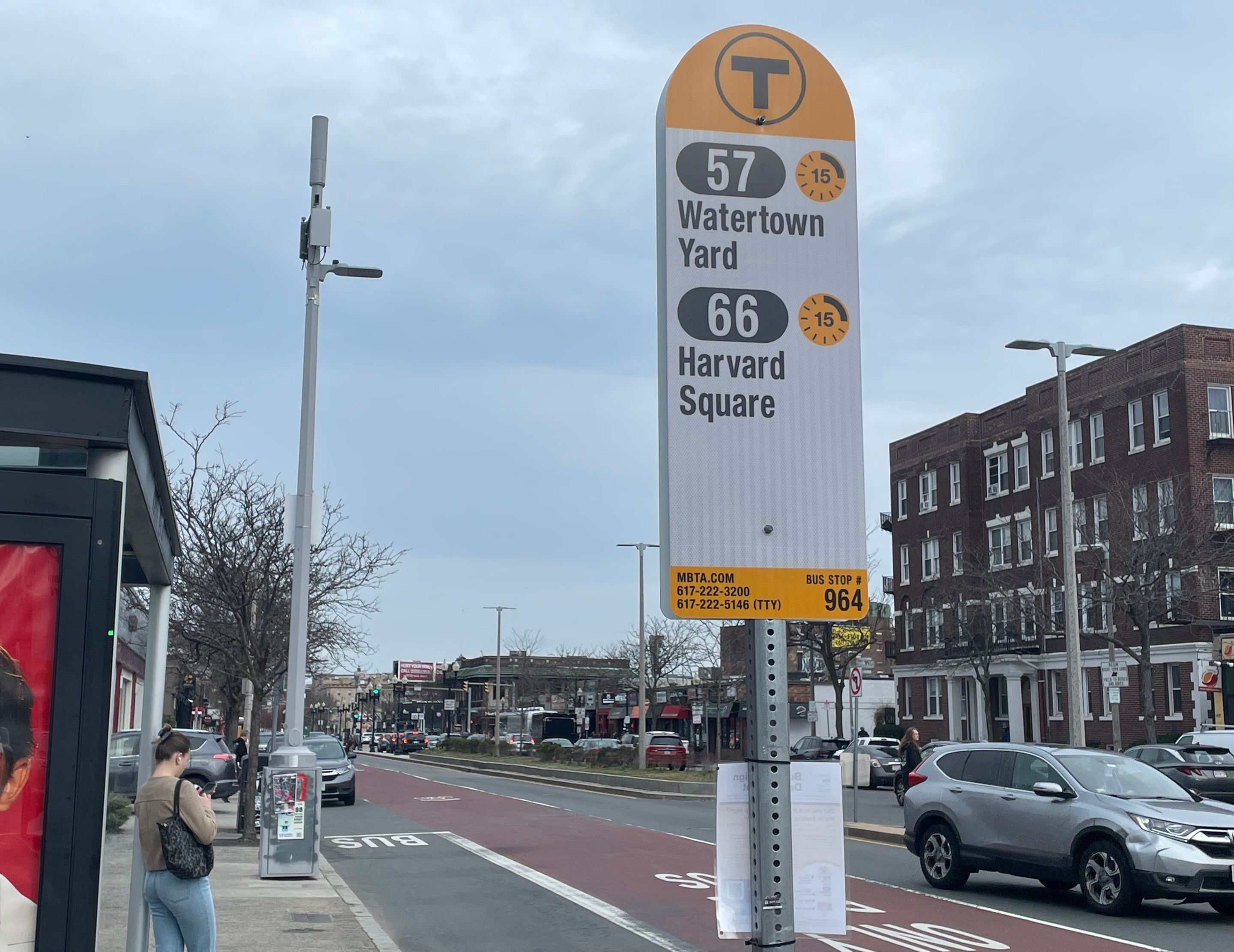 A bus stop sign next to a bus shelter where a passenger is waiting shows two bus routes - the 75 to Watertown Yard and the 66 to Harvard Square - next to a yellow clock icon with a "15" in the middle.