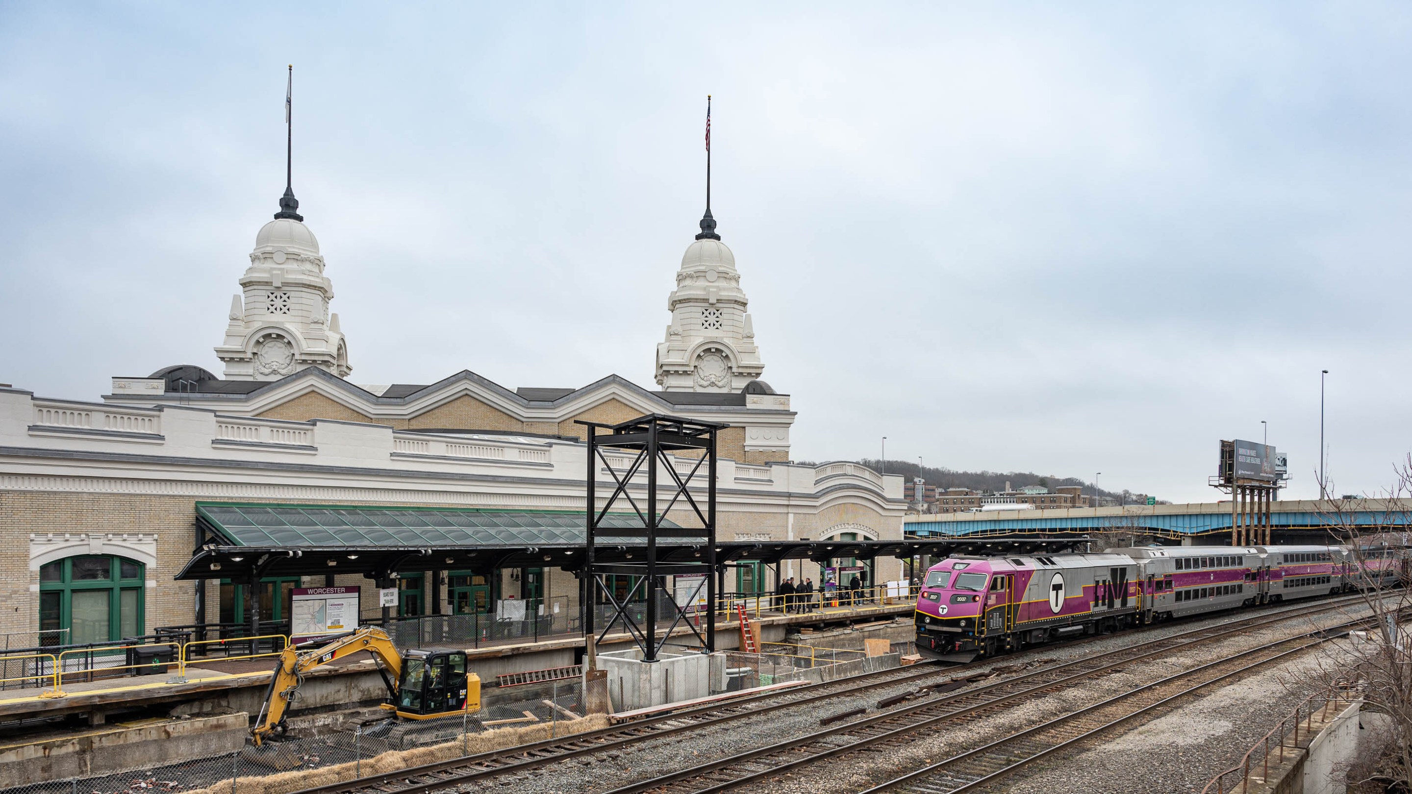 An MBTA commuter rail train with a purple front and a T logo on its side approaches an under-construction station platform in the center of the image. In the right foreground are two other railroad tracks. To the left behind the platform are two domed spires rising above a train station building.