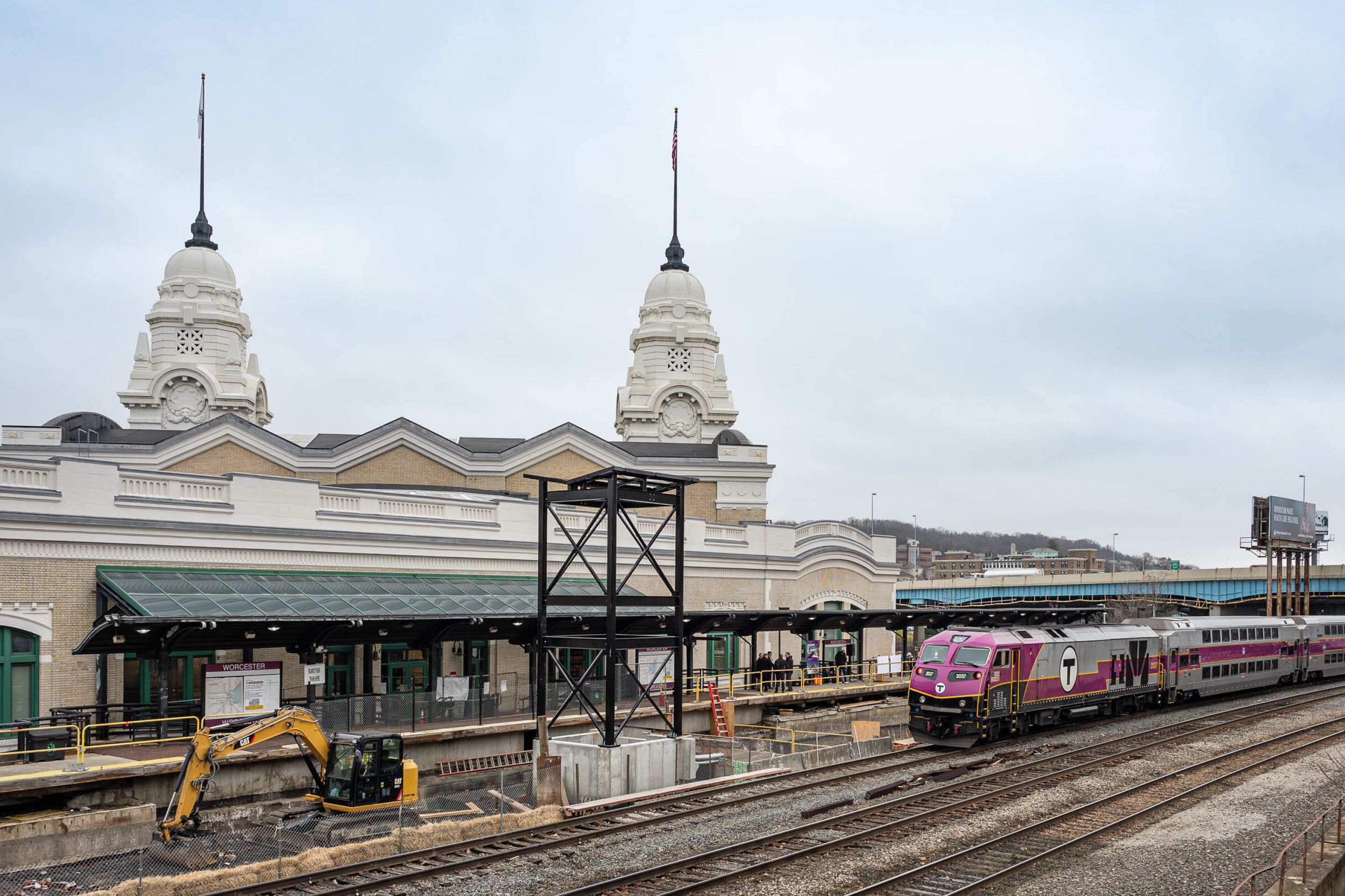 An MBTA commuter rail train with a purple front and a T logo on its side approaches an under-construction station platform in the center of the image. In the right foreground are two other railroad tracks. To the left behind the platform are two domed spires rising above a train station building.