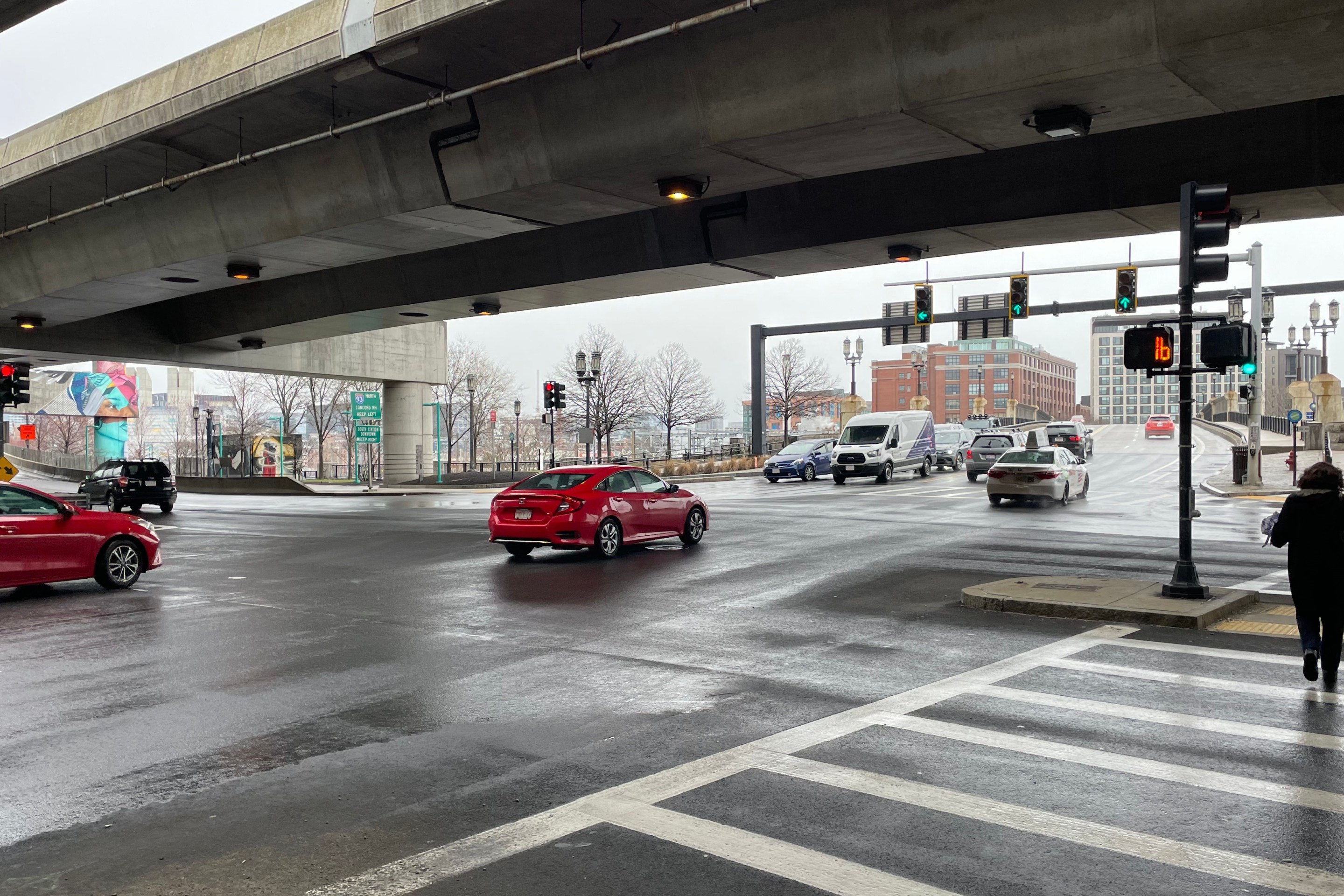 Several cars speed through a wide multi-lane intersection underneath a concrete highway viaduct. On the left edge of the photo are signs for I-90 and I-93. On the right edge, a woman crosses several lanes of traffic in a crosswalk.