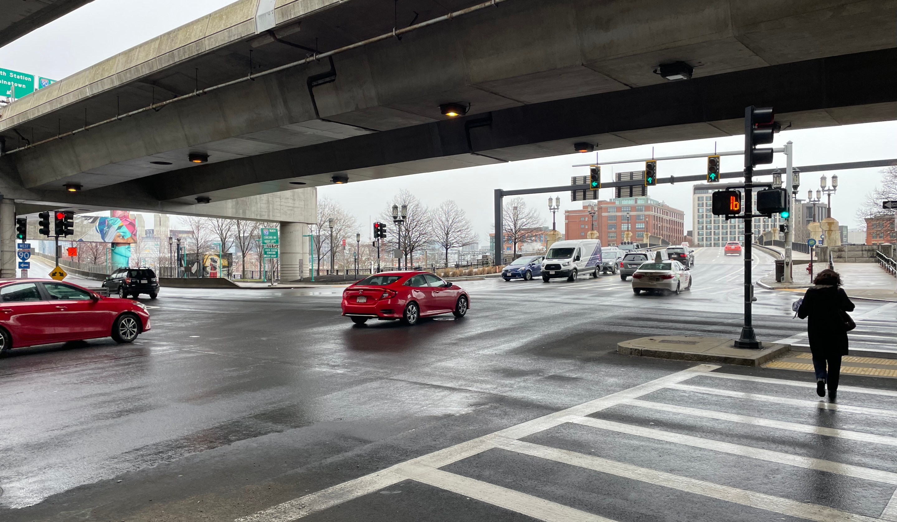 Several cars speed through a wide multi-lane intersection underneath a concrete highway viaduct. On the left edge of the photo are signs for I-90 and I-93. On the right edge, a woman crosses several lanes of traffic in a crosswalk.