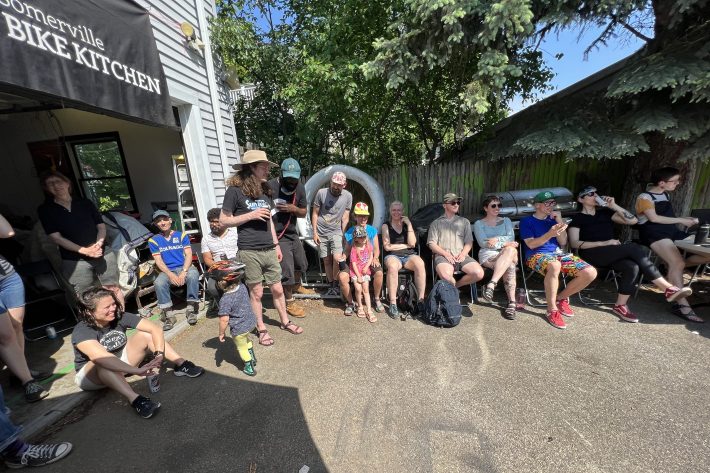 Spectators wearing shorts and t-shirts sit around the edge of a paved driveway in front of an open garage door with a banner above it that reads "Somerville Bike Kitchen"