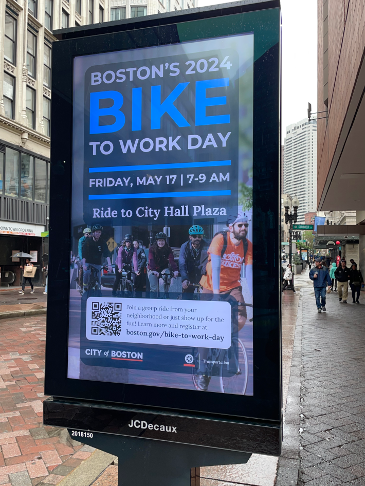 An electronic ad sign on the street in Downtown Crossing displays an ad for "Boston's Bike to Work Day Friday May 17 7-9 AM - Ride to City Hall Plaza" above a photo of 6-7 people riding their bikes on a city street.