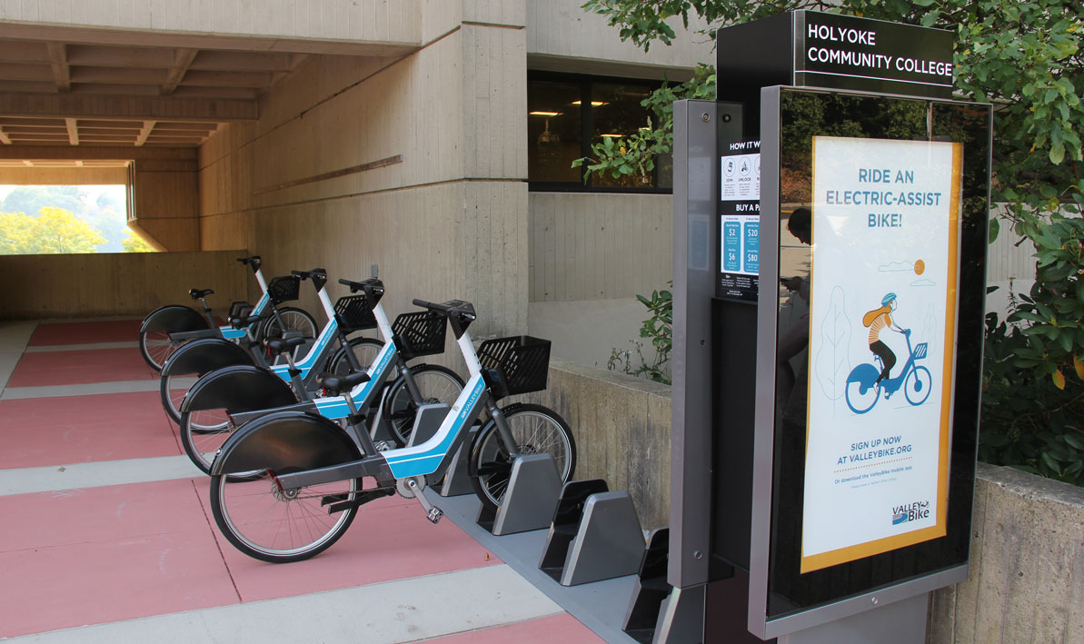 A bikesharing station kiosk with four parked bikes in a sheltered passageway of a concrete building. The kiosk has a sign that says "Holyoke Community College" and an ad that says "ride an electric-assist bike"