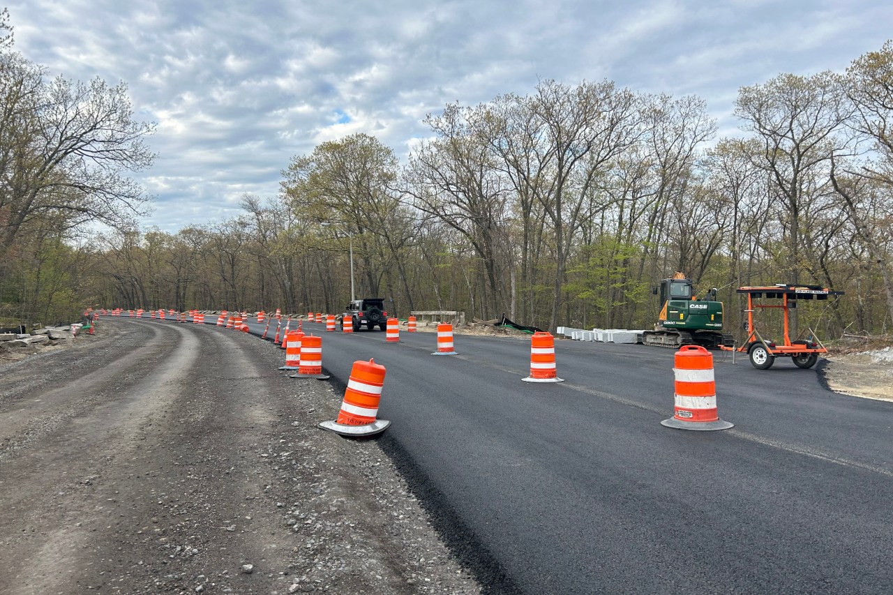 A photo of a street construction site running through woods that are just beginning to leaf out with spring leaves. On the left side of the photo is a roughly 20-foot-wide expanse of dirt and gravel. To the right is a freshly-paved two-lane road, lined with orange construction barrels. On the far side of the pavement is an excavator and an orange construction trailer.