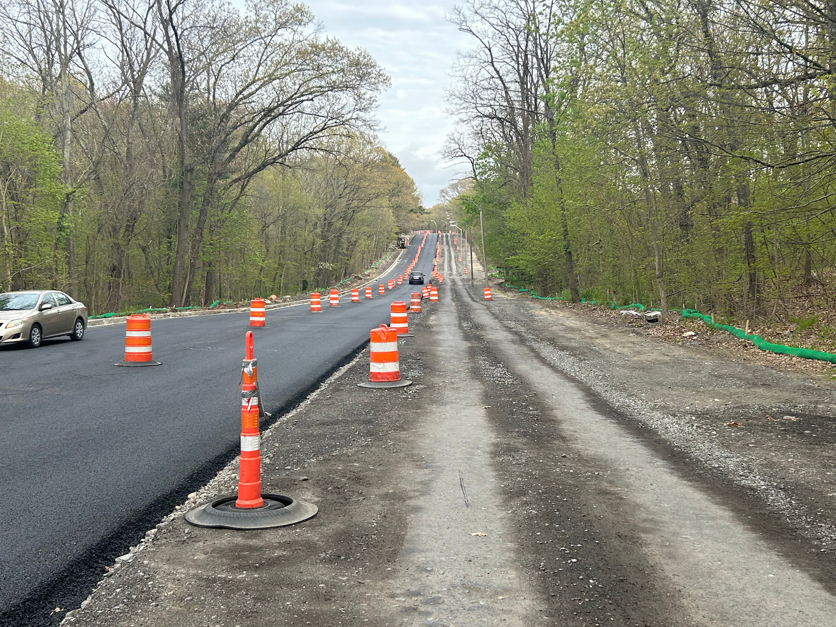 A freshly-paved two-lane road is lined with orange construction barrels stretching into the distance. The road is surrounded by trees just beginning to grow their leaves. A wide dirt track runs along the right edge of the pavement.