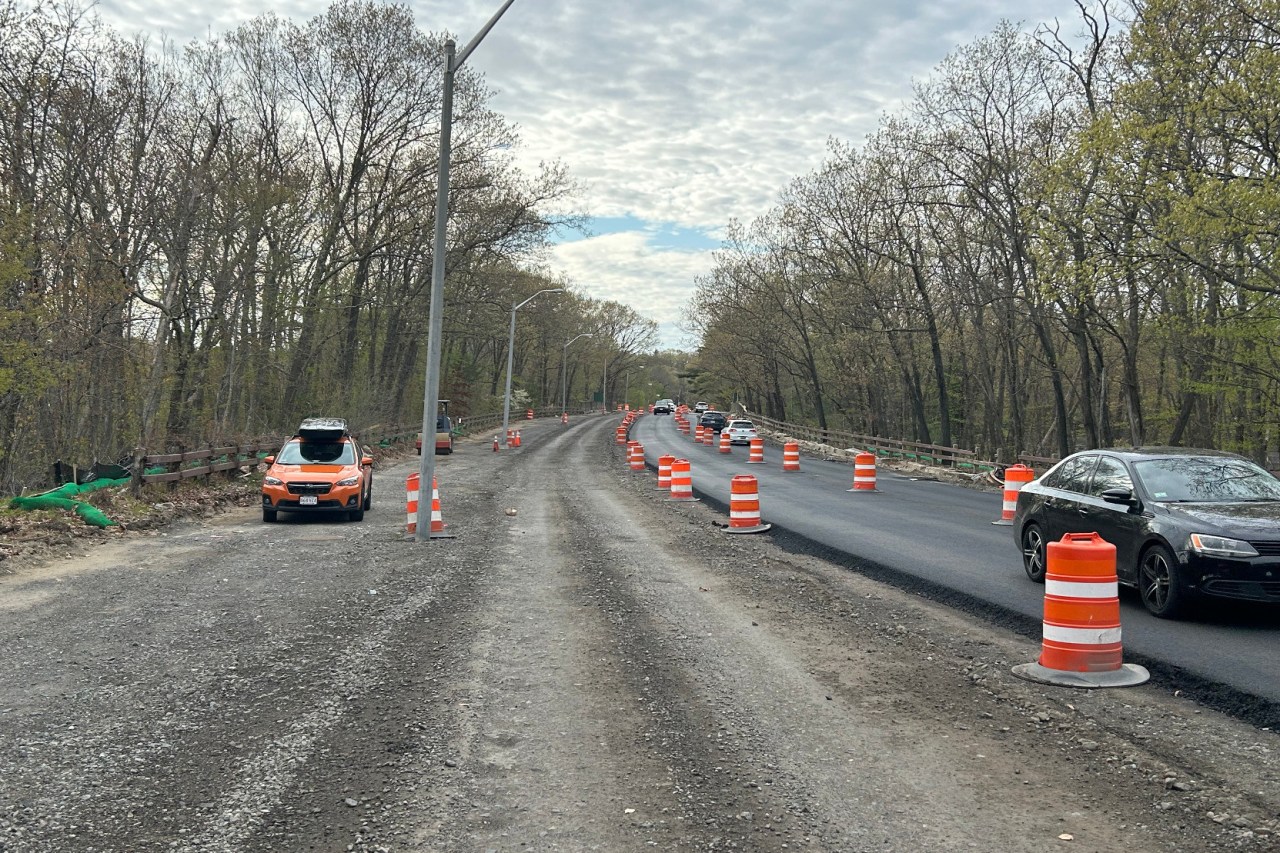 A dirt track runs alongside a freshly-paved two-lane road on the right side of the photo. The edge of the pavement is lined with orange construction barrels. The road is surrounded by trees just beginning to grow their leaves.