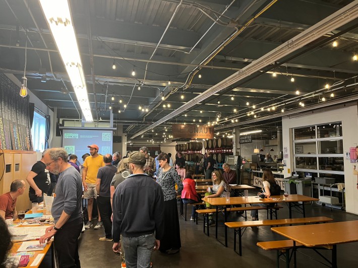 People gathered at Aeronaut Brewing. Large hall with exposed ceiling and string lights across it, picnic tables with people sitting and talking, people standing around bike organizations' tables, talking to tabling staff and observing materials on the table.