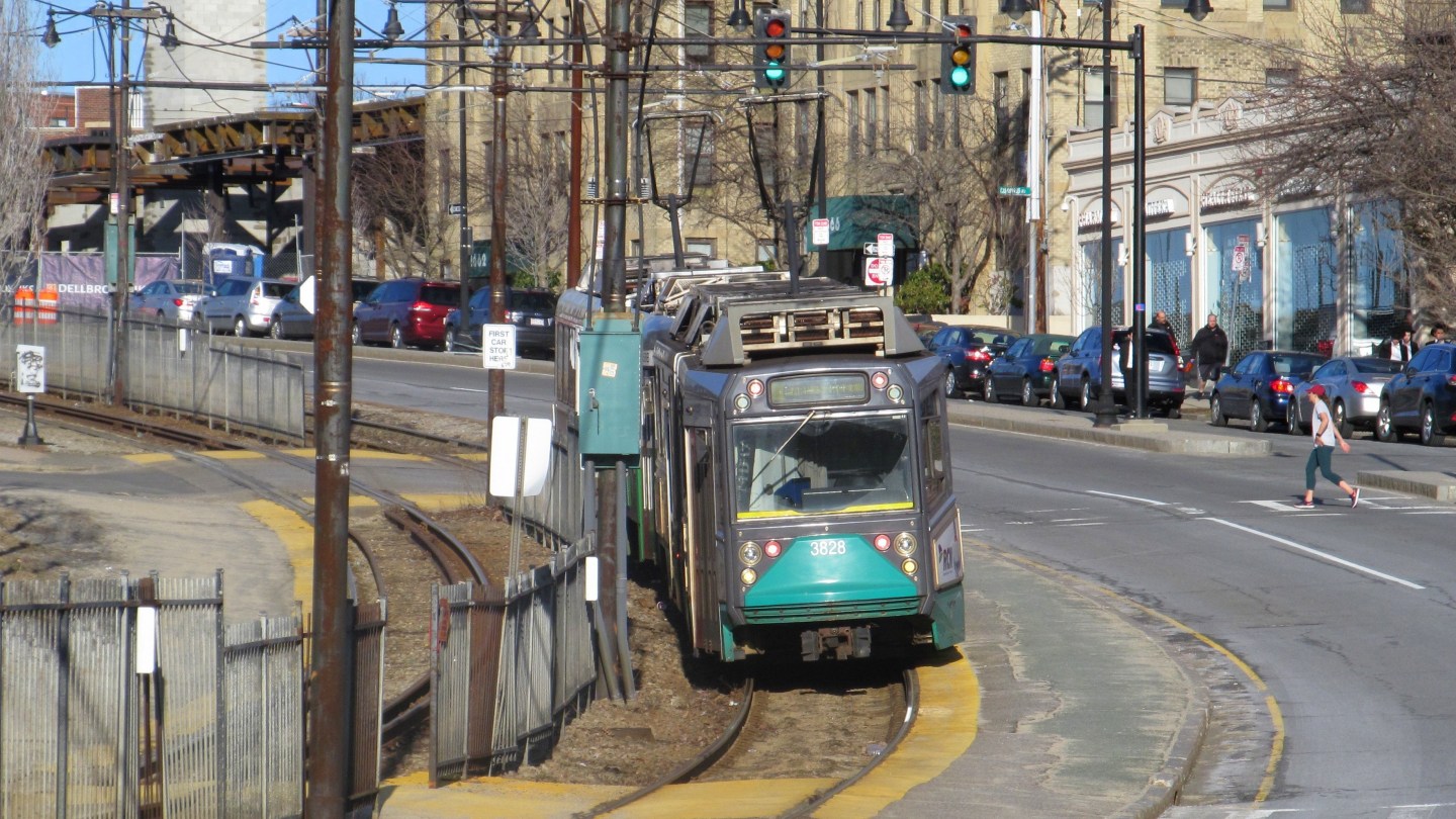 A green line train stops in the median of a multi-lane roadway as a pedestrian crosses in a crosswalk towards it.