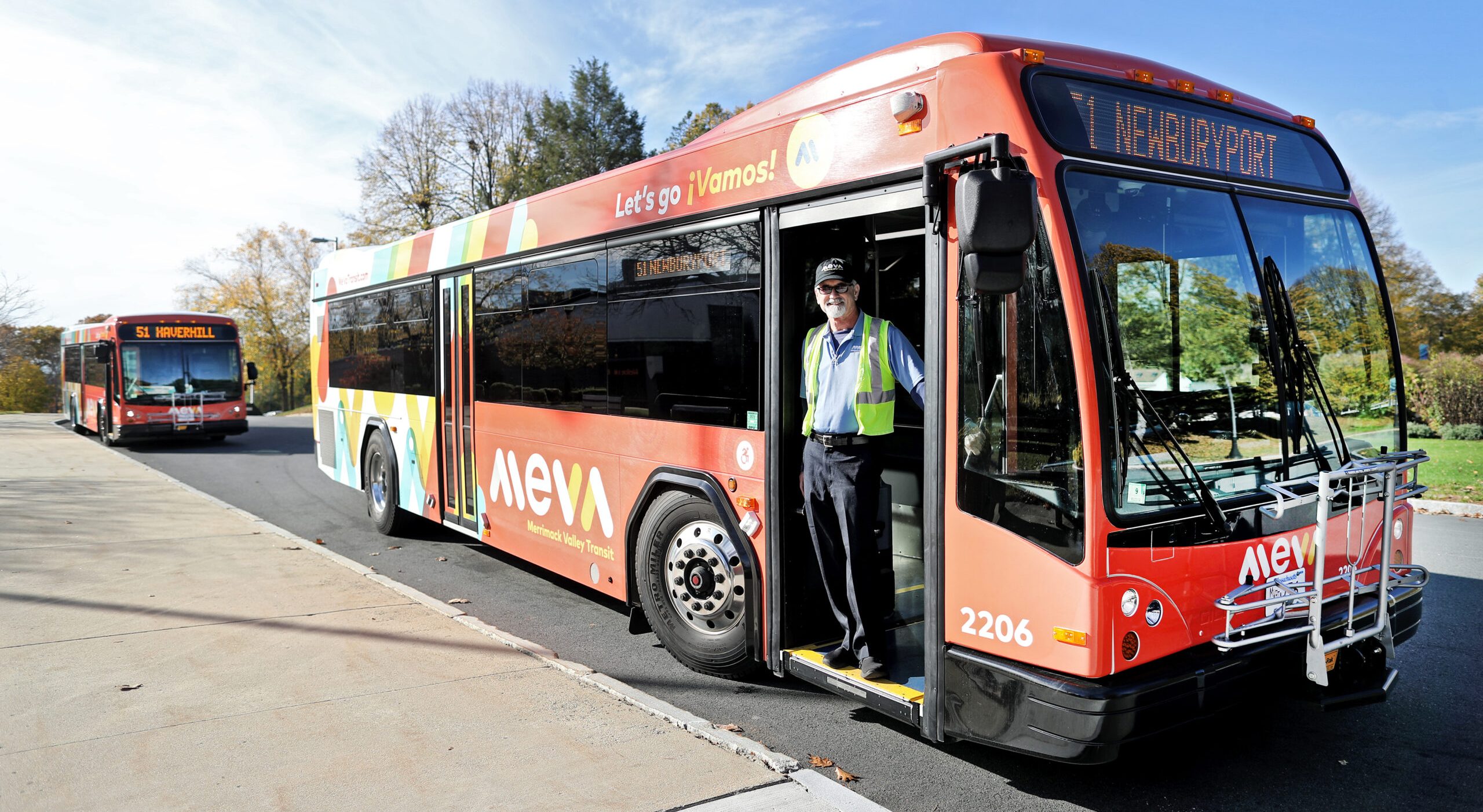 A driver with a white beard wearing a green vest and a black baseball cap stands in the door of a red city bus with the MeVa logo on its side. The destination sign on the front of the bus says "61- Newburyport" and a second bus waits behind in the background at left.