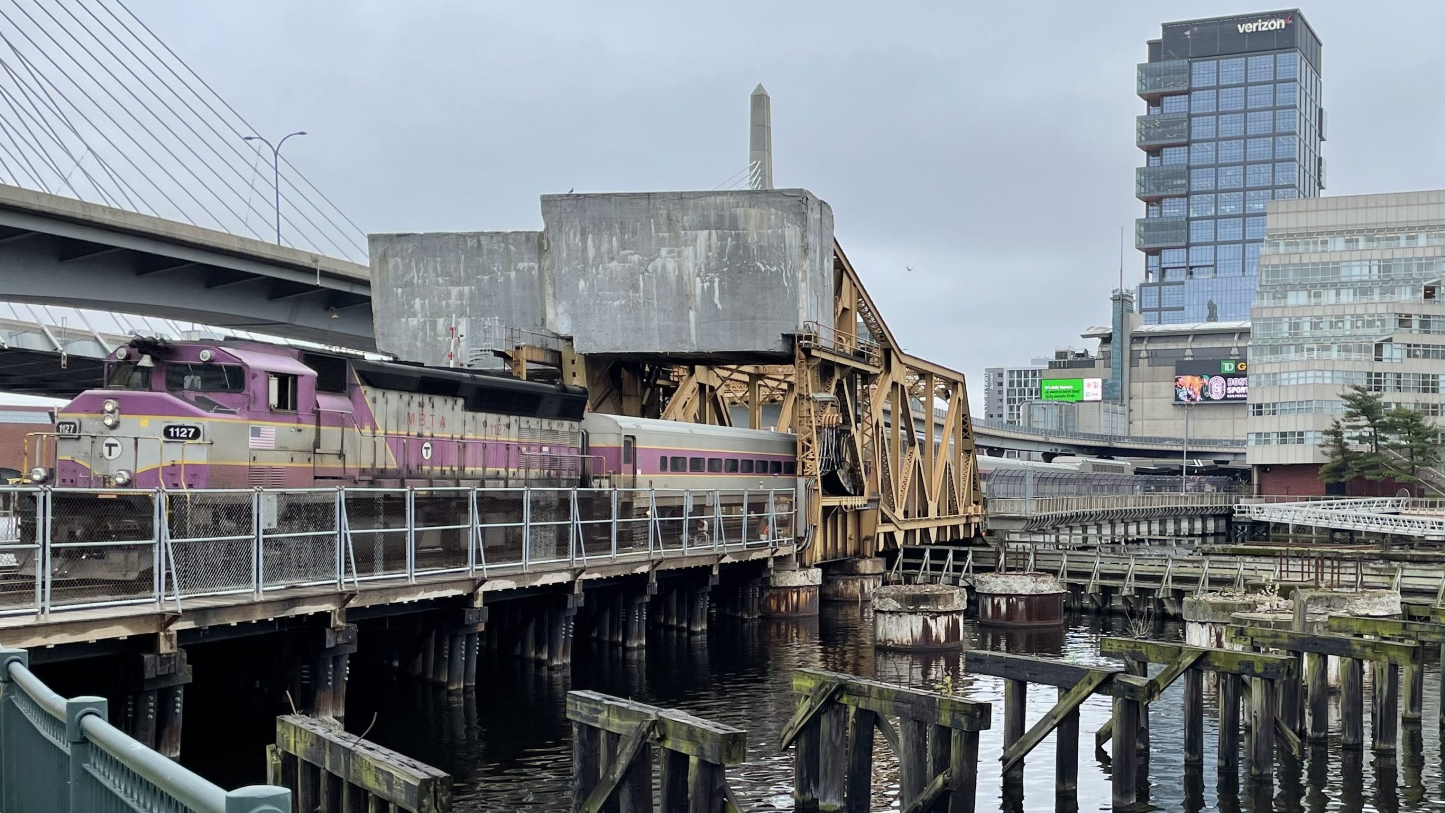 A purple-and-silver MBTA commuter rail diesel train crosses the North Station drawbridge over the Charles River with the high-rises of downtown Boston in the background.