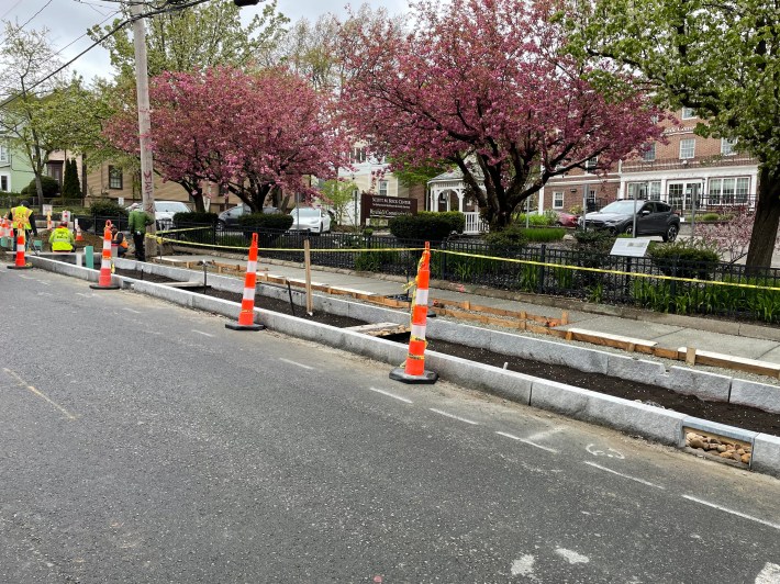 Several orange construction cones protect granite curbs that delineate a rectangular planting bed along the edge of a city street. At the far left edge of the photo several workers in fluorescent yellow vests are working. Beyond the edge of the sidewalk is a parking lot lined with several trees blooming with pink blossoms.