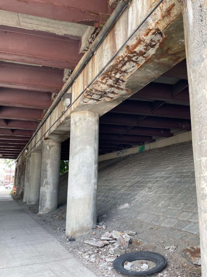 The underside of a highway overpass with a concrete beam held up by columns. The bottom of the beam shows extensive deterioration with rusted metal rebar exposed, and chunks of fallen concrete line the sidewalk below.