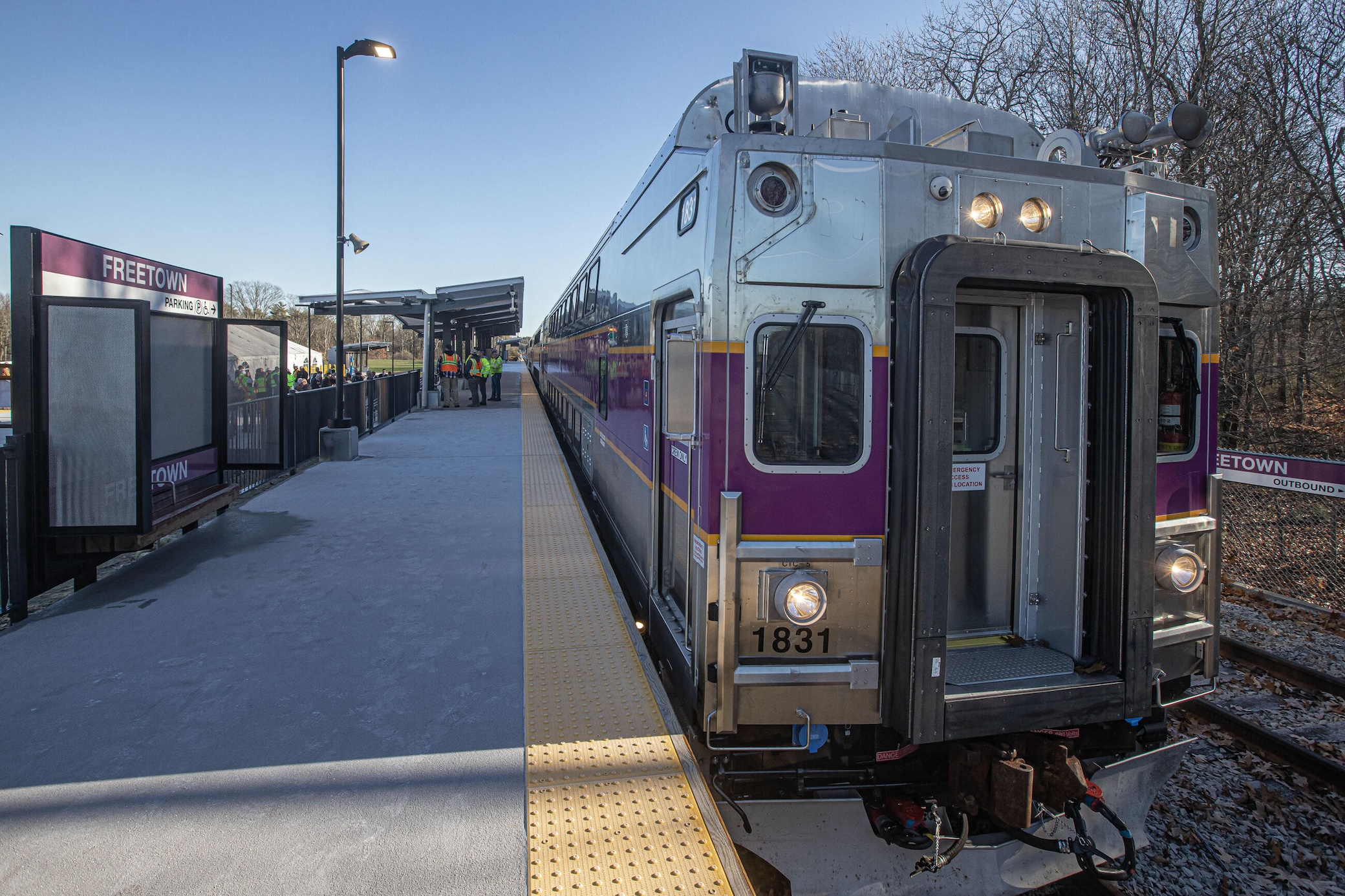 The end of an MBTA commuter rail train next to a train platform. Purple signs on the platform indicate it is Freetown station. Several workers in yellow vests are under a canopy at the far end of the platform in the distance.