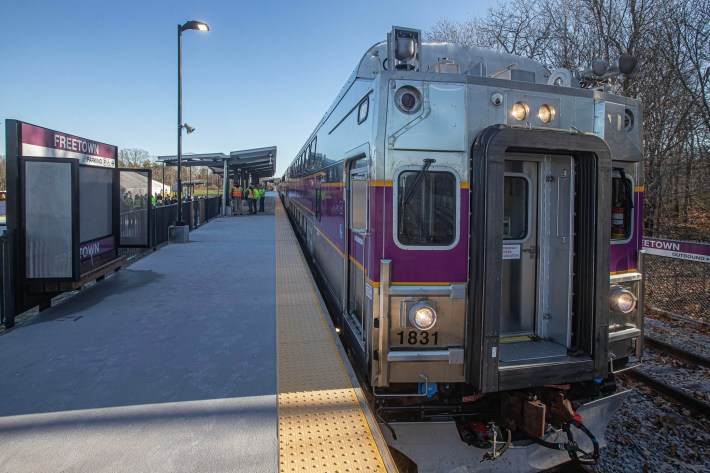 The end of an MBTA commuter rail train next to a train platform. Purple signs on the platform indicate it is Freetown station. Several workers in yellow vests are under a canopy at the far end of the platform in the distance.