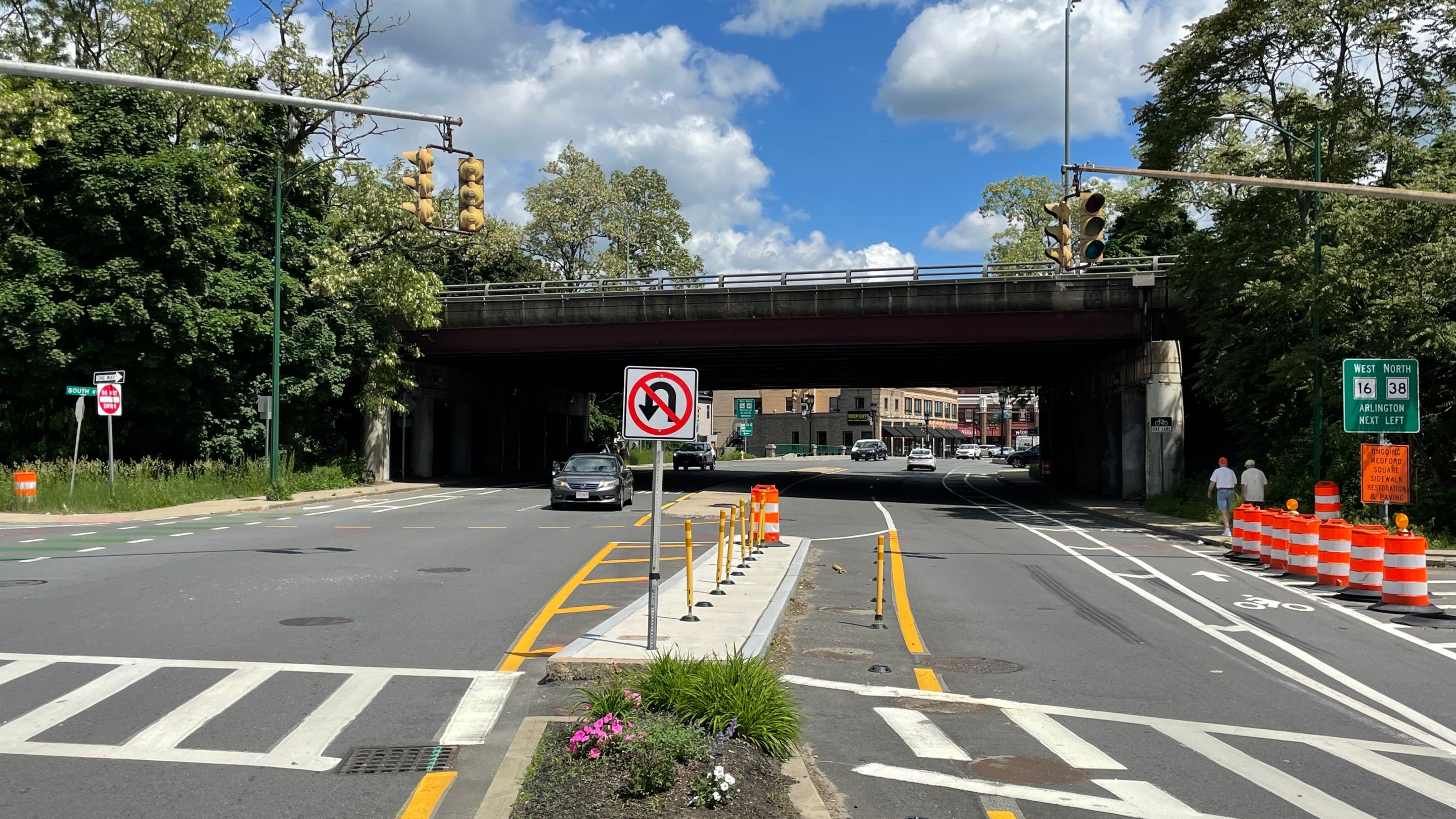 A view of a wide, multi-lane street leading under two traffic signals with a dark highway overpass beyond.