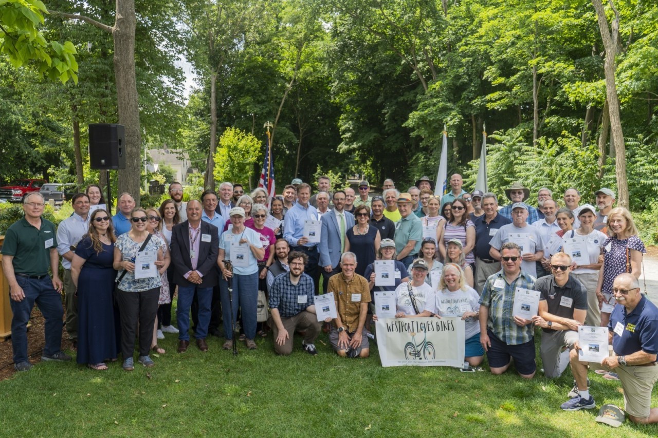 A crowd of people on a grassy lawn surrounded by trees smiles for the camera while holding up certificates.