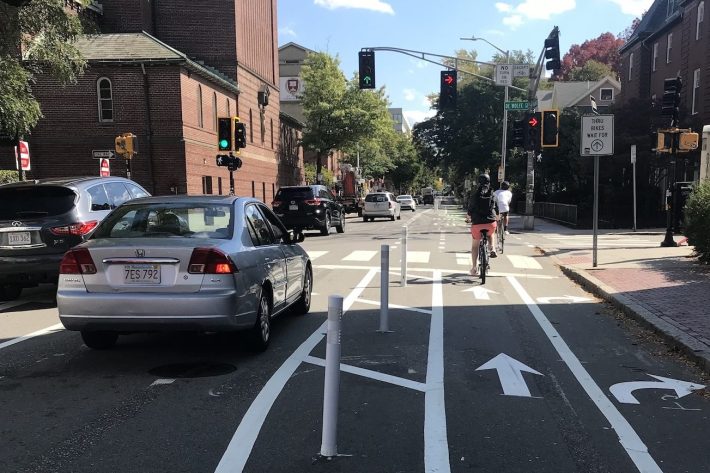 A street with flexible post bollards to divide the car lanes (left) from the bike lanes (right). The bikeway has a right-turn lane against the curb next to a lane with straight arrows for through bike traffic. The street is lined with old brick buildings.