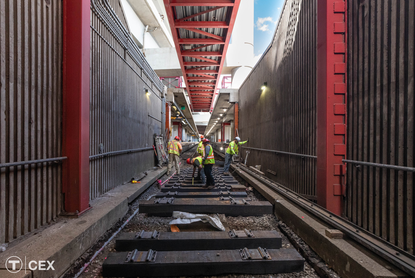 Workers replace rails in a subway tunnel, with a station platform visible in the distance.