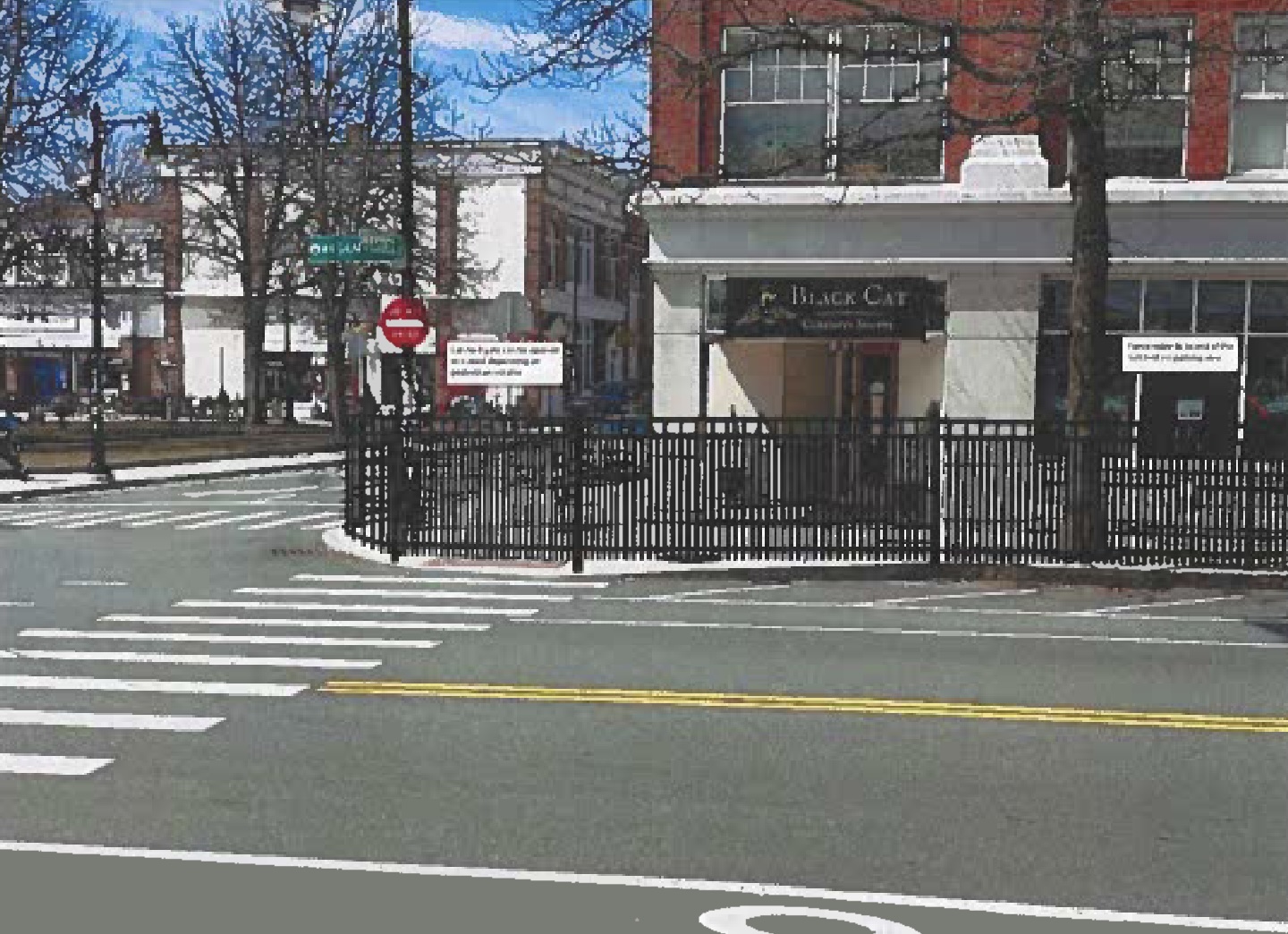 A rendering of a city street with a black metal fence surrounding the sidewalk on the far side of the street. In the background is a brick multi-story building