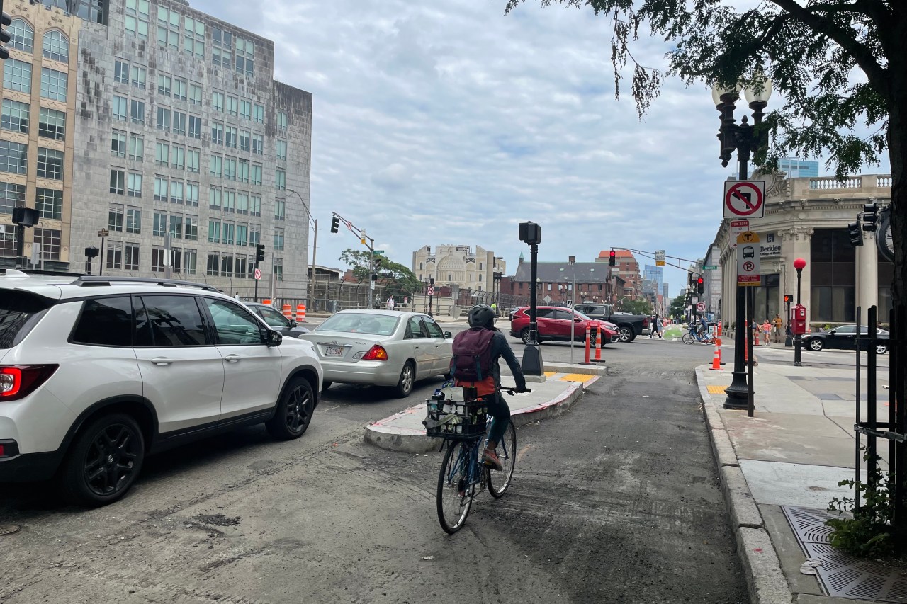 A person on a bike approaches an intersection in a bike lane that's separated from the adjacent car lane by a granite curbed island with a walk signal. The street is surrounded by high-rise buildings and there are several orange construction cones in the intersection.