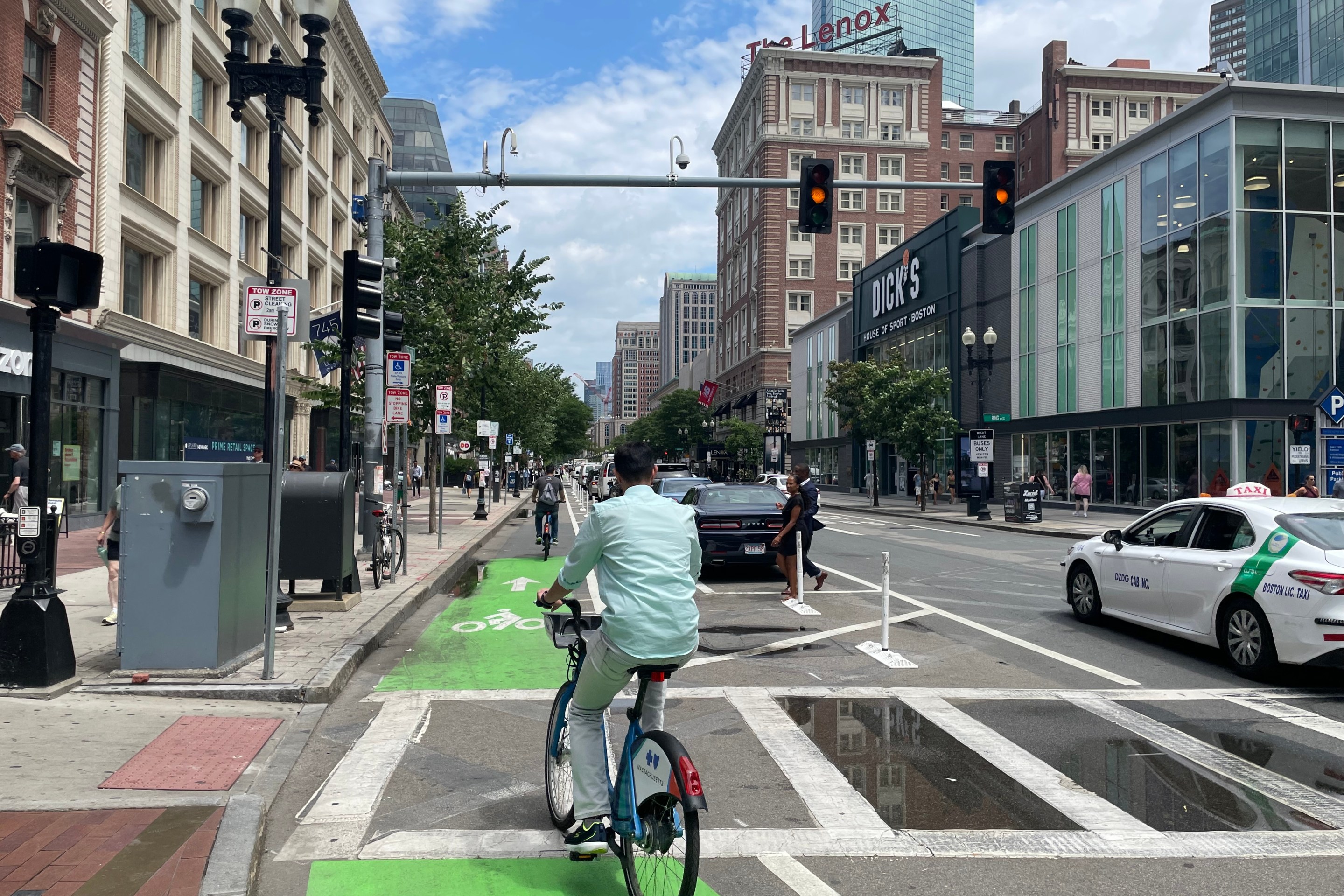 A person in a teal dress shirt rides through a crosswalk on a green bike lane that lies between a row of parked cars in the middle of the street and a busy sidewalk on the left.