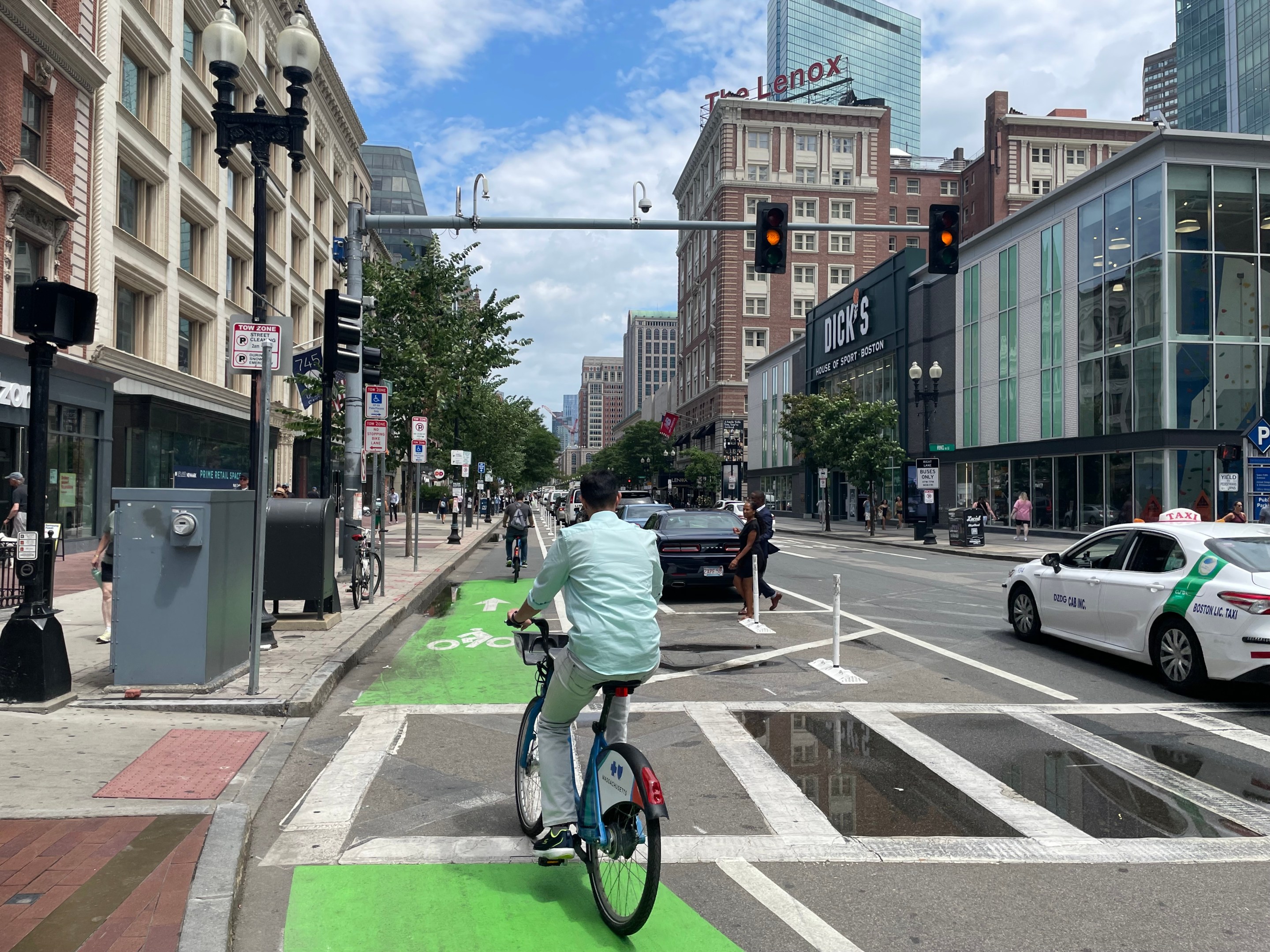 A person in a teal dress shirt rides through a crosswalk on a green bike lane that lies between a row of parked cars in the middle of the street and a busy sidewalk on the left.