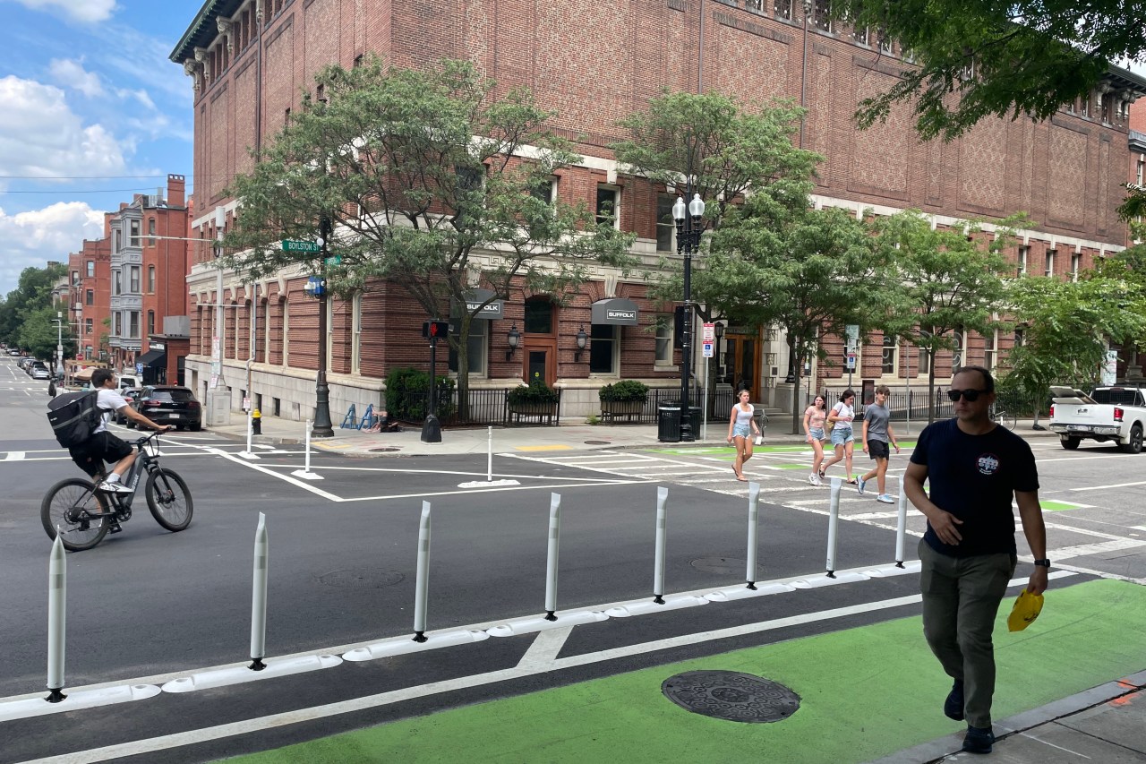 A person on a bike and five pedestrians cross a street lined with flexible-post bollards that separate a green bike lane in the foreground from motor vehicle lanes in the middle of the street. In the background is a large brick building.