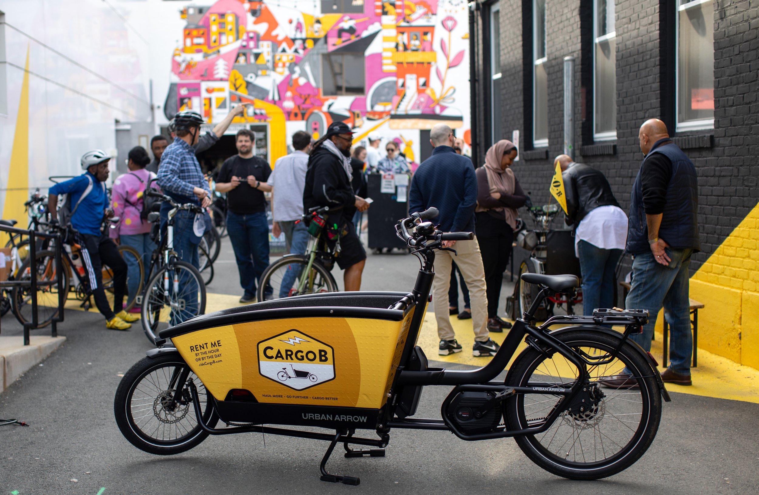 A CargoB cargo bike with a bright yellow bucket in front of the handlebars in the alley outside Aeronaut brewery with a brightly colored mural in the background