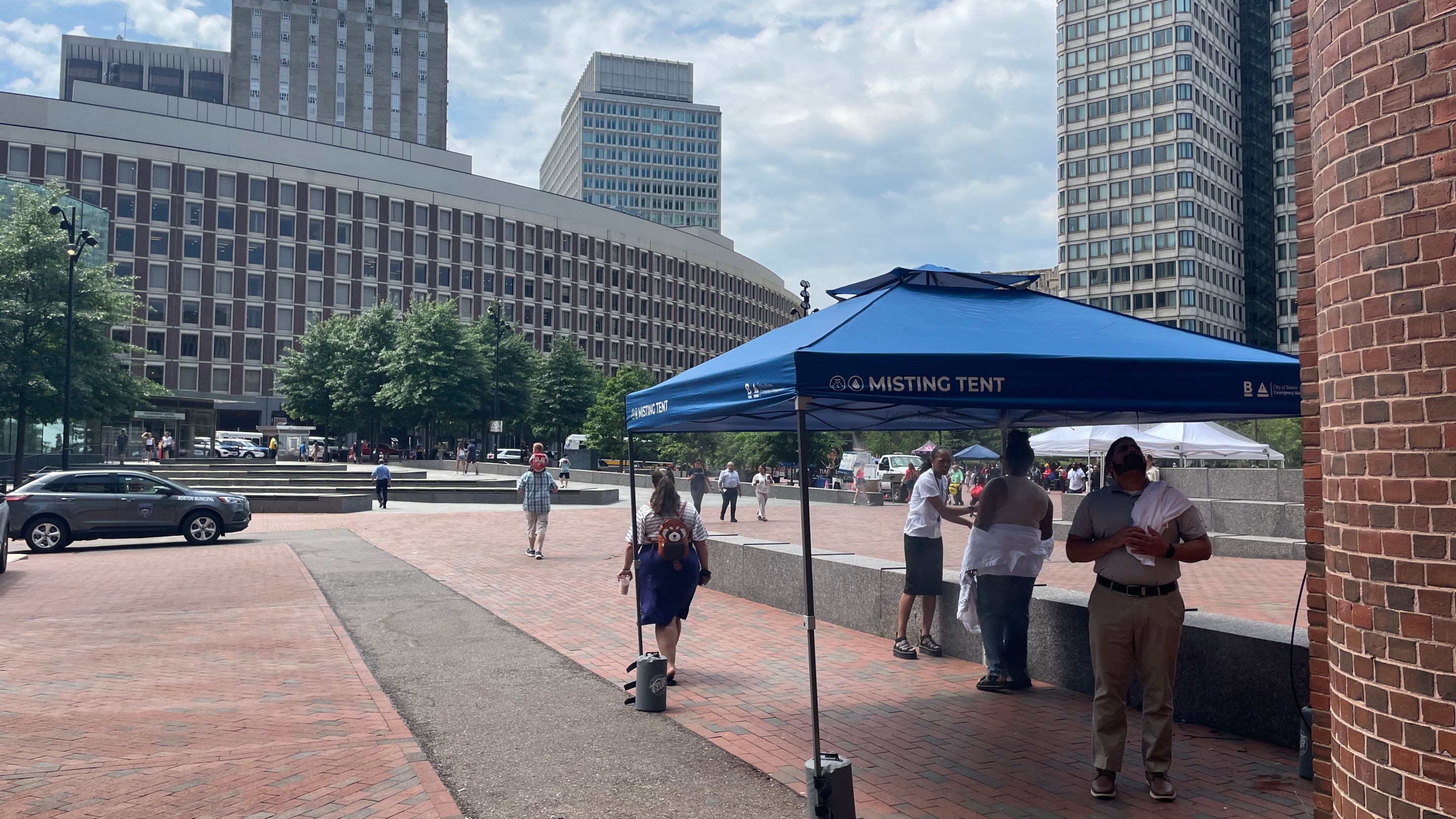 Four people gather under a blue pop-up tent labelled "misting station" on a wide open brick plaza on a sunny day, with high-rise buildings in the distance.